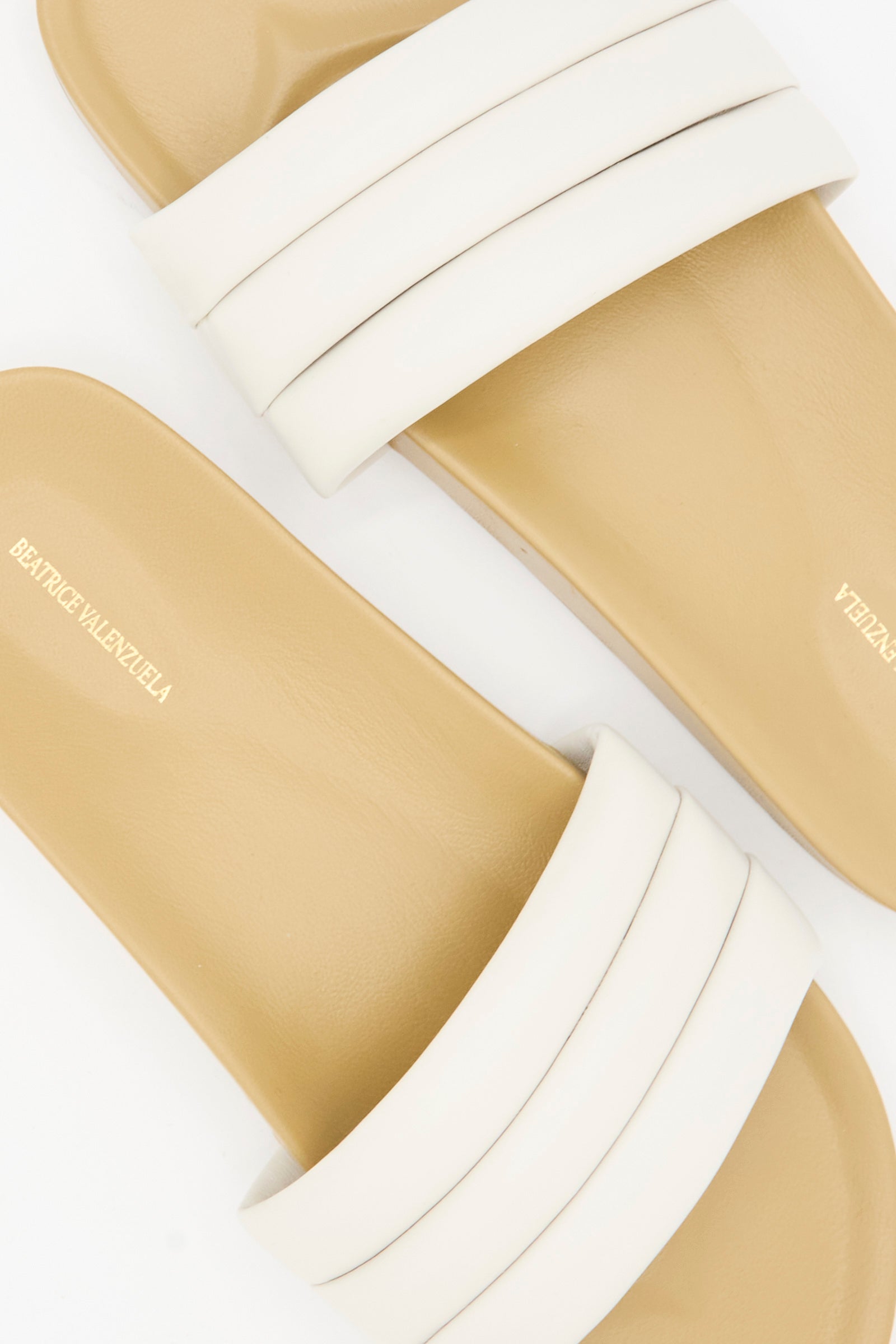 The Classic Sandalia in Shell by Beatrice Valenzuela features tan and white handcrafted slide sandals with two wide straps and the brand name printed on the insole.