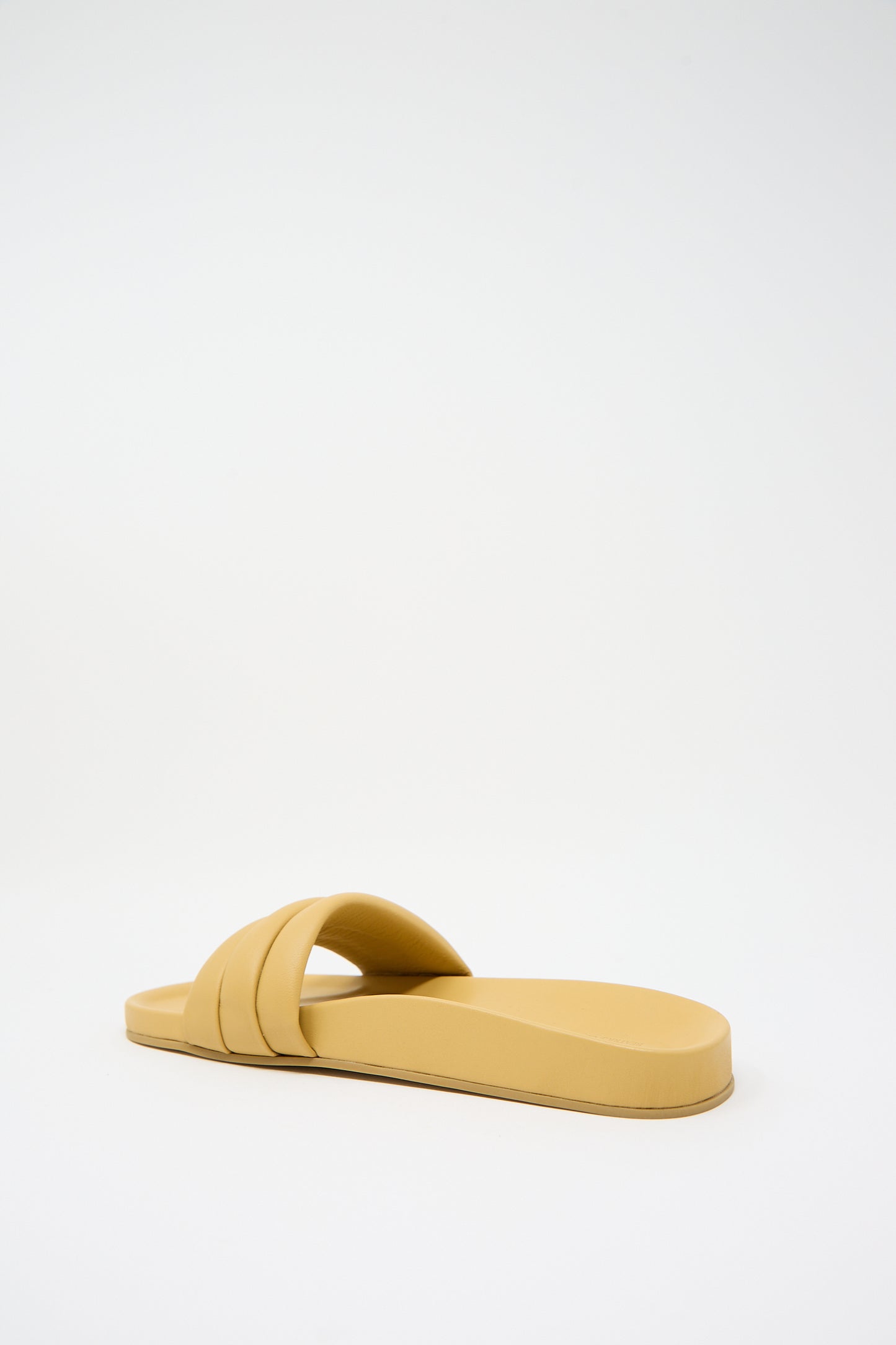 A single, beige-colored slip-on sandal with a double strap design made from luxurious lambskin uppers rests on a plain white background. It is the Monocolor Sandalia in Wheat by Beatrice Valenzuela.