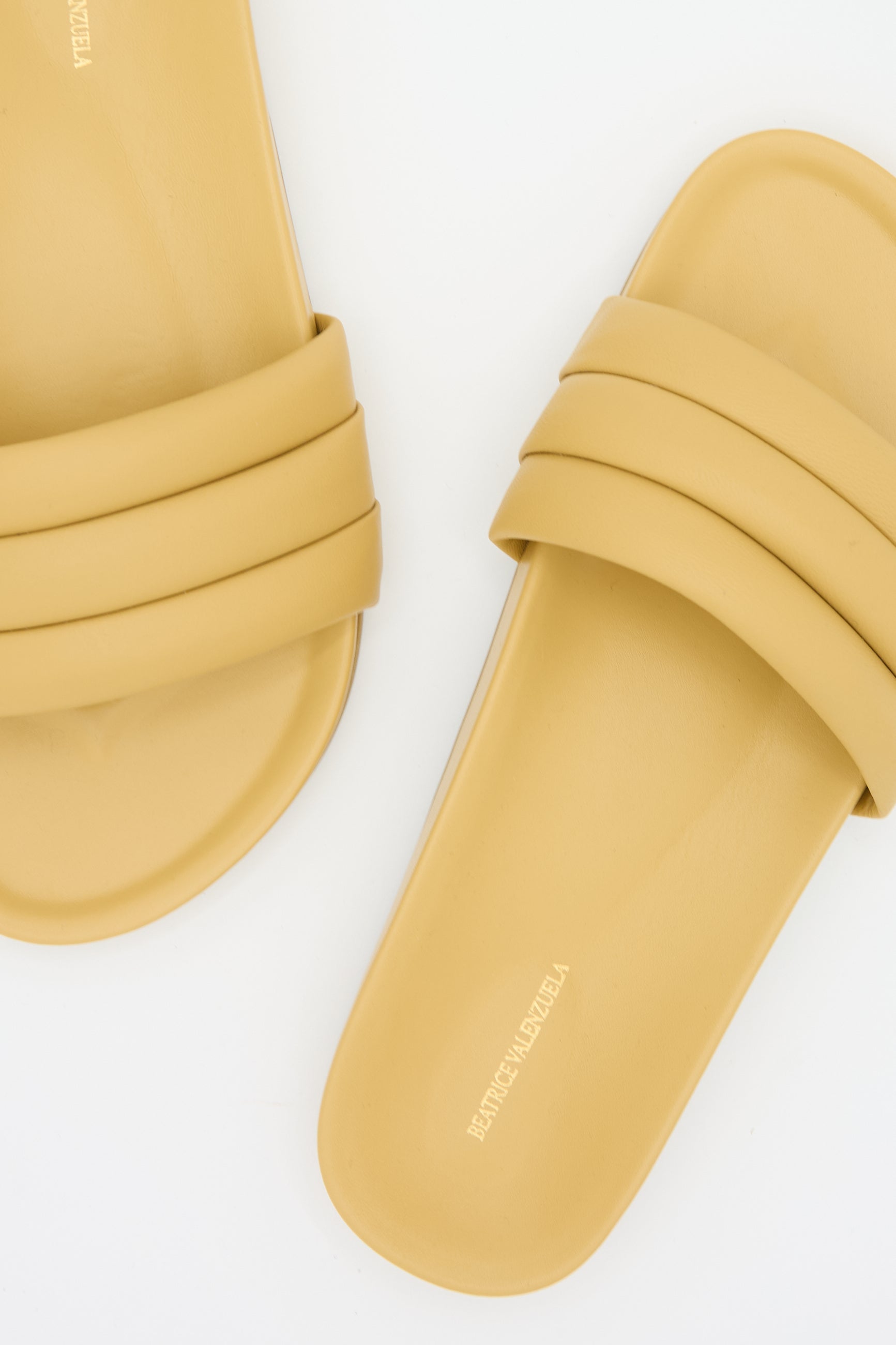 Two Monocolor Sandalia in Wheat by Beatrice Valenzuela with double straps, positioned on a white background. These handcrafted slides feature lambskin uppers, and the brand name is embossed on the insole.