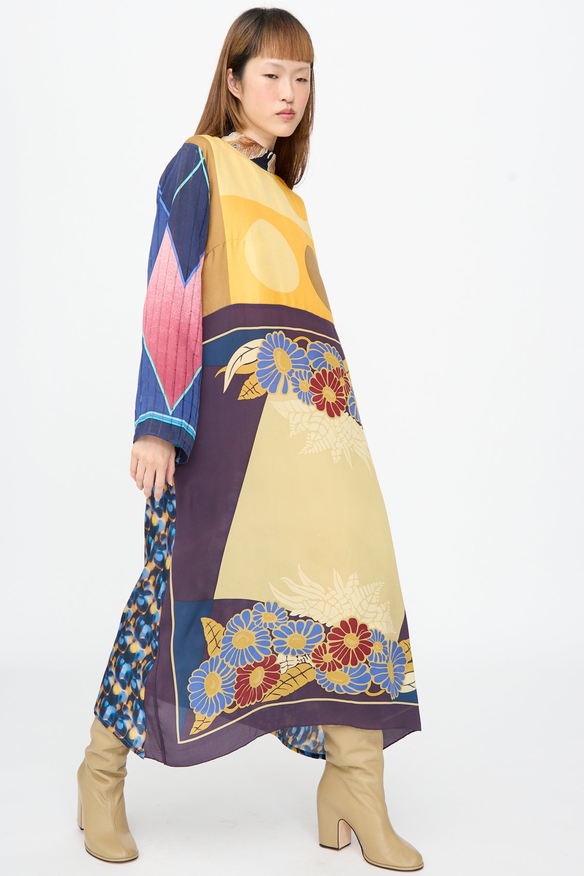 A model wears the Bettina Bakdal Classic Mandarin Collar Dress in Alice Vollmer, a long-sleeve midi dress with a patchwork of blue, red, and yellow floral and geometric shapes, styled with tan heeled boots.