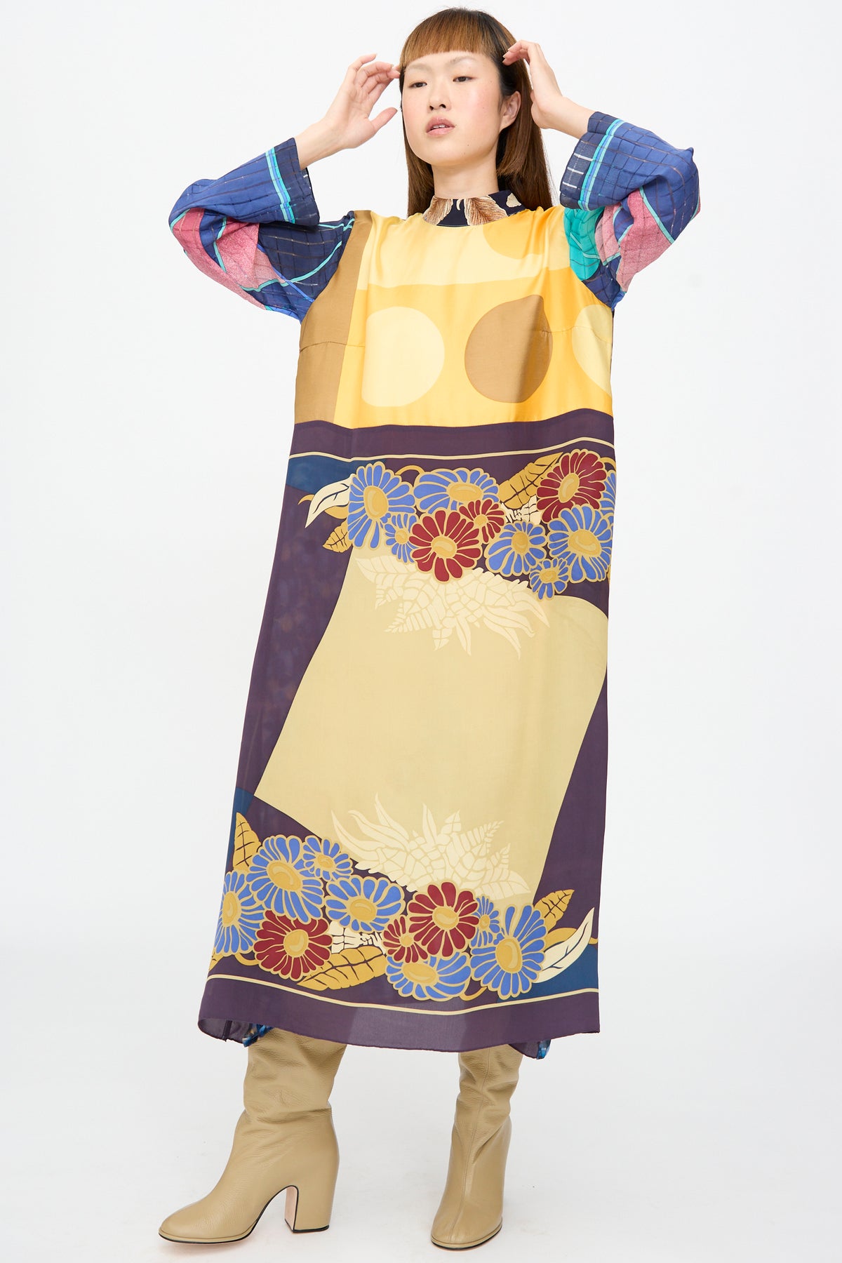 A person wearing the Bettina Bakdal Classic Mandarin Collar Dress in Alice Vollmer, a long-sleeved, knee-length up-cycled silk dress in a yellow, purple, and blue floral pattern, with beige ankle boots.