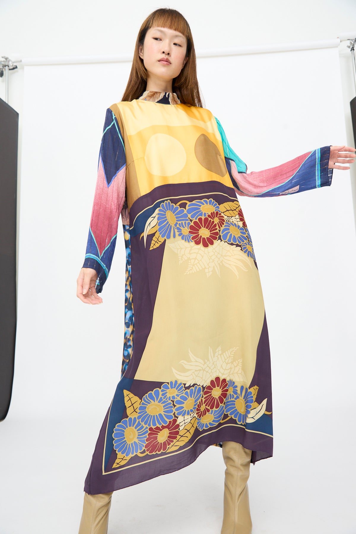 A person wearing a Bettina Bakdal Classic Mandarin Collar Dress in Alice Vollmer, a knee-length, long-sleeved up-cycled silk dress with a multicolored floral and geometric print, paired with tan boots.