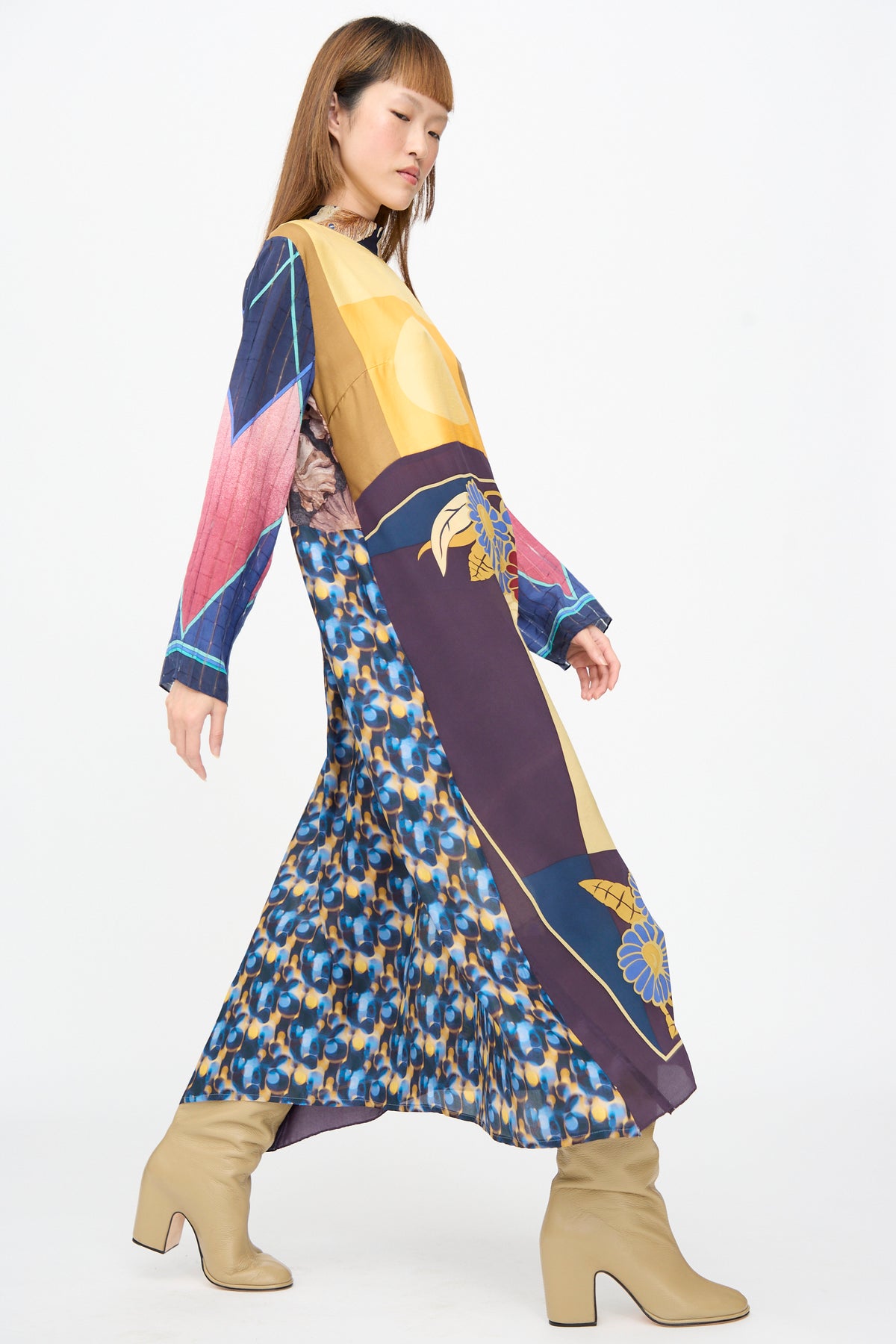 A person models the Bettina Bakdal Classic Mandarin Collar Dress in Alice Vollmer, a knee-length, long-sleeve dress made from colorful vintage silk scarves, paired with tan block-heeled boots.
