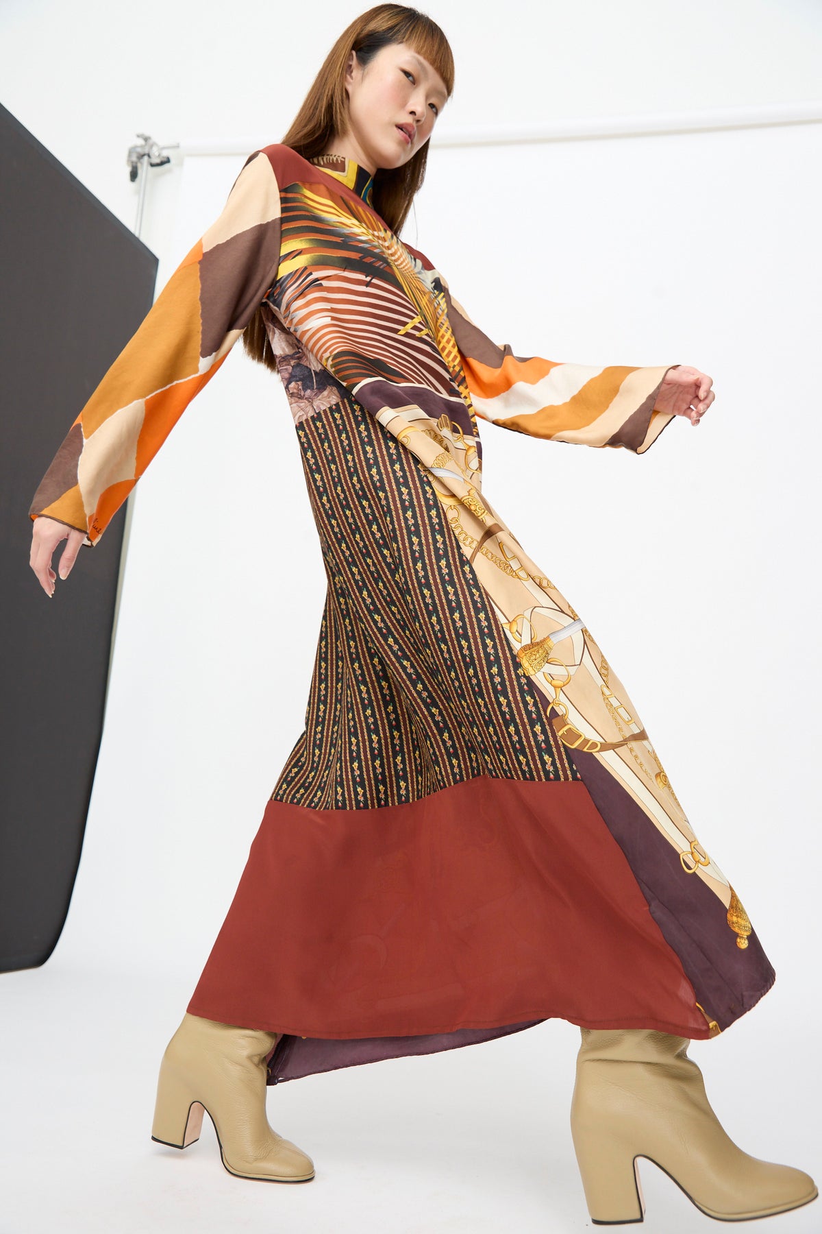 A person wearing a Bettina Bakdal Classic Mandarin Collar Dress in Benita Otte, featuring a long-sleeved up-cycled silk midi silhouette with a colorful geometric and chain-link pattern and tan block-heeled boots.
