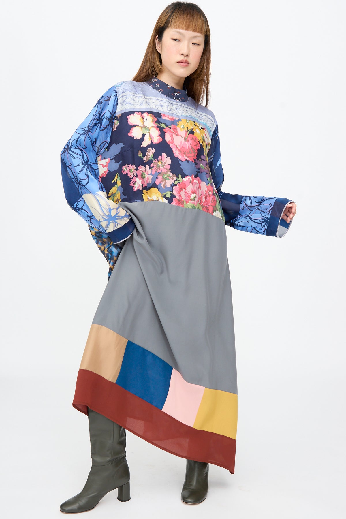 A model wears the Bettina Bakdal Classic Mandarin Collar Dress in Grete Willers, an up-cycled silk midi featuring a patchwork floral and blue design, grey skirt with color-blocked hem, and olive boots.