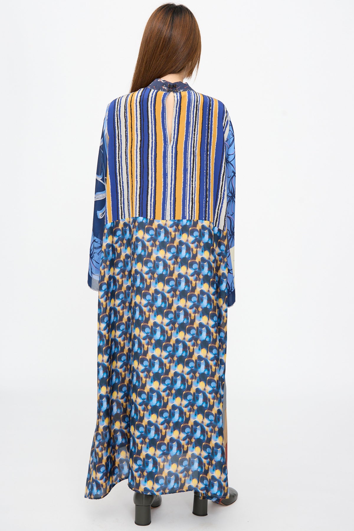Back view of a woman in a Bettina Bakdal Classic Mandarin Collar Dress in Grete Willers, a long-sleeve, floor-length silk dress with a patchwork of blue, yellow, and white geometric and striped patterns.