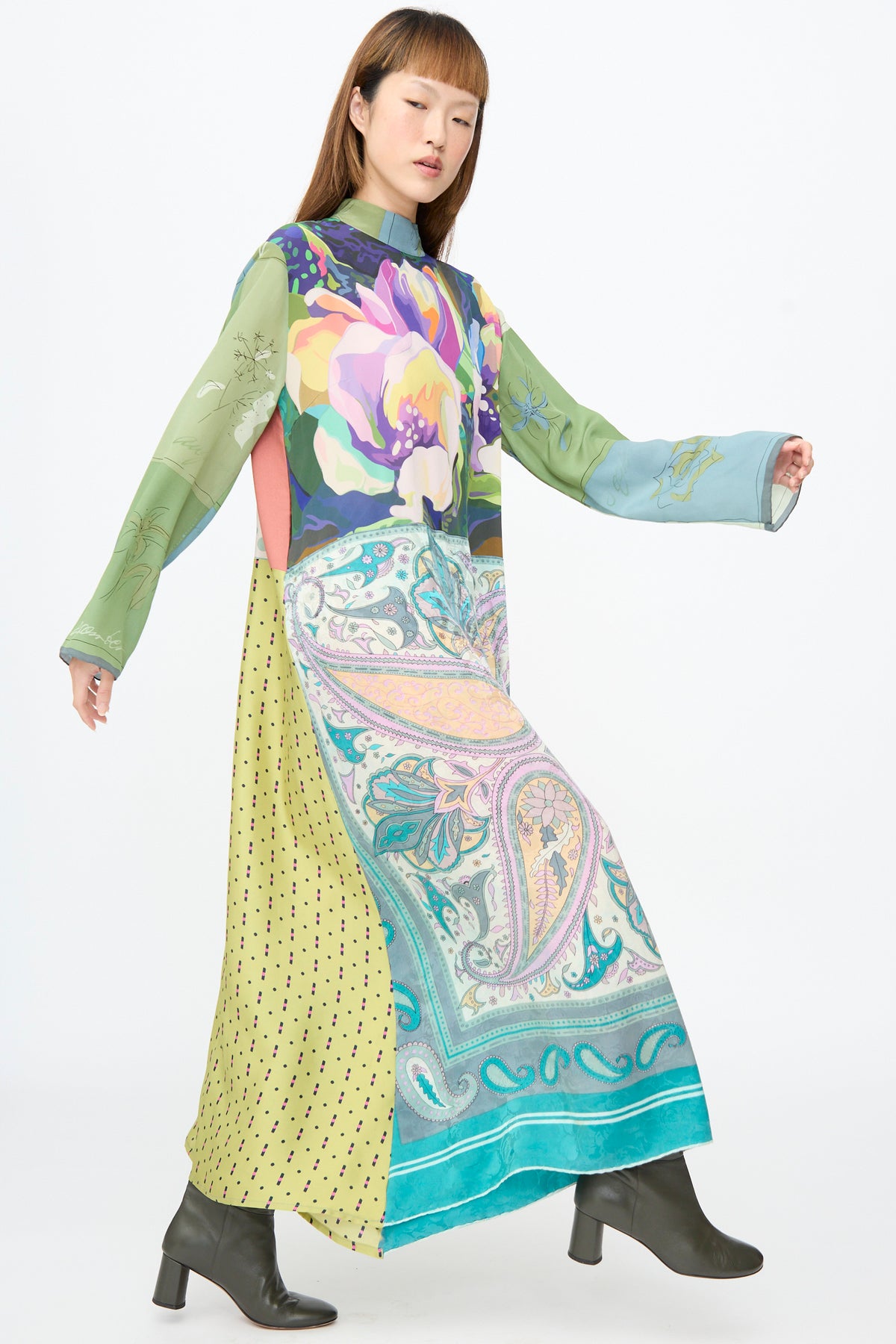 A person wears a Bettina Bakdal Classic Mandarin Collar Dress in Hedwig Arheim, a long-sleeved, calf-length up-cycled silk dress in green and teal floral, paisley, and polka-dot patterns, with dark boots.