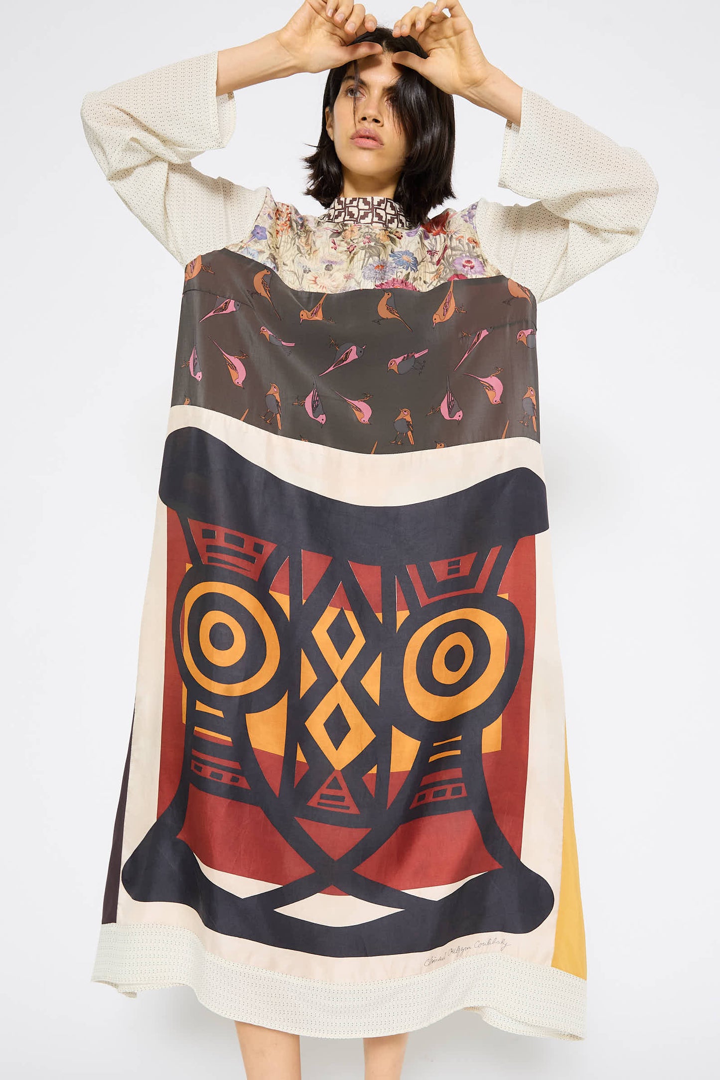 A person stands holding a woven item with a large colorful geometric pattern featuring eyes and bold shapes, elegantly complementing their Classic Mandarin Collar Dress in Little Pink Songbird by Bettina Bakdal.