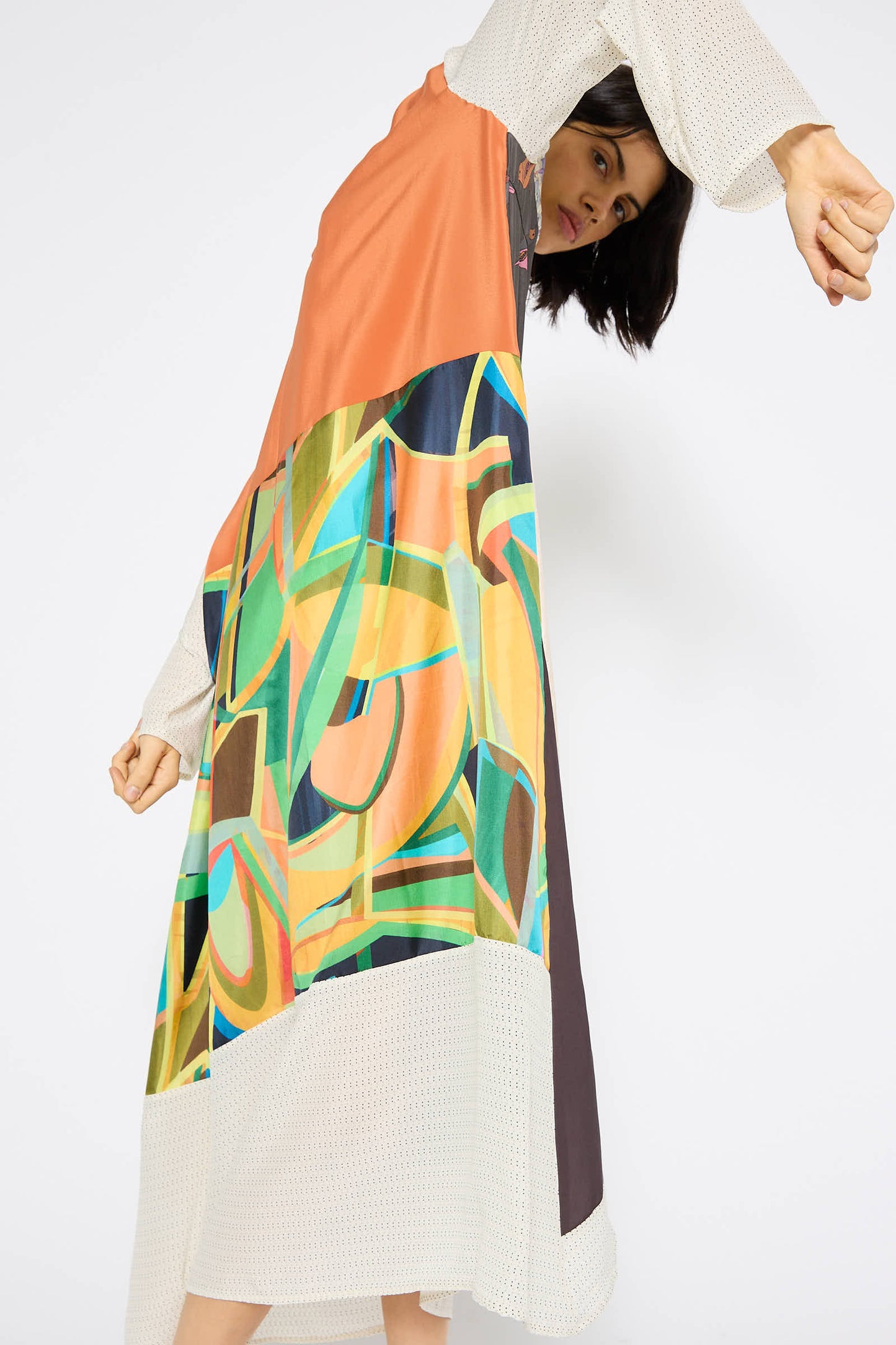 A person wearing the Bettina Bakdal Classic Mandarin Collar Dress in Little Pink Songbird, featuring a vibrant geometric pattern and crafted from up-cycled vintage silk scarves, poses with one arm extended and head tilted against a plain background.