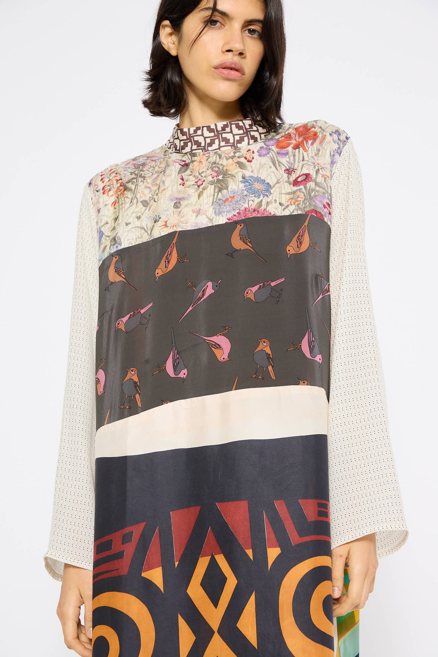 A person stands against a plain background wearing the "Classic Mandarin Collar Dress in Little Pink Songbird" by Bettina Bakdal, fashioned from up-cycled vintage silk scarves featuring floral, bird, and geometric designs.