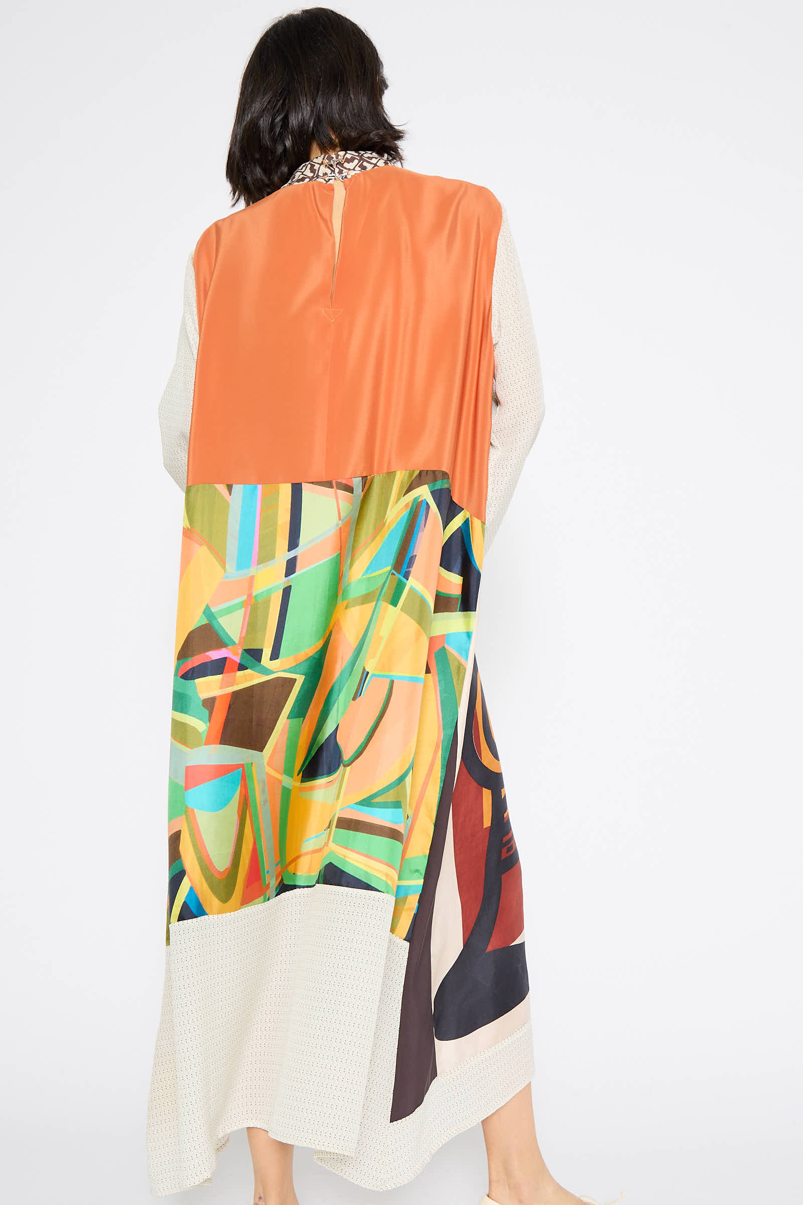 A woman with shoulder-length hair wears the Classic Mandarin Collar Dress in Little Pink Songbird by Bettina Bakdal, made from up-cycled vintage silk scarves and featuring geometric patterns in shades of orange, green, and cream, as she is viewed from behind.