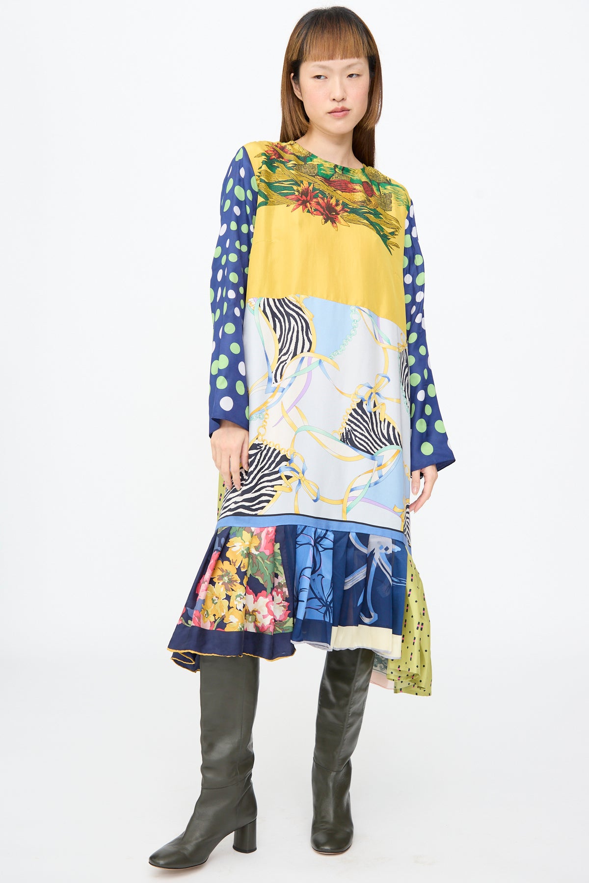 A person wearing the Bettina Bakdal Hannah Dress in Friederike Dicker, a knee-length patchwork dress with yellow florals, blue polka dots, zebra print, and chain-link motifs, paired with olive boots.