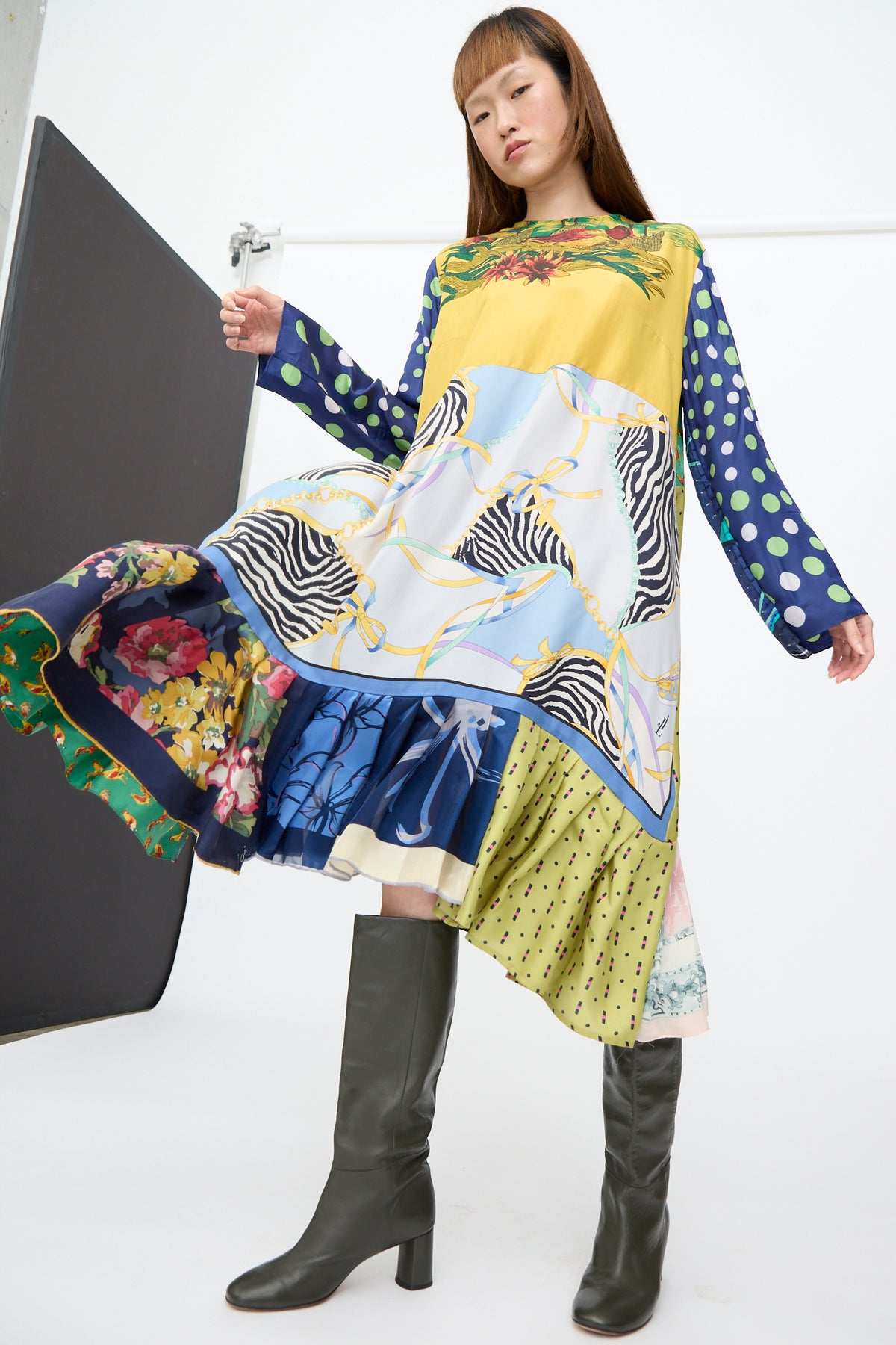 A person wears a knee-length Hannah Dress in Friederike Dicker featuring floral, polka dot, and chain-link patterns, paired with dark green knee-high boots.
