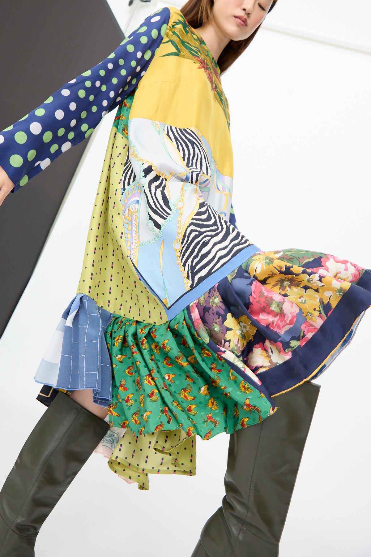 A person wearing the Bettina Bakdal Hannah Dress in Friederike Dicker, a long, multicolored patchwork print dress featuring floral, polka-dot, and zebra patterns, paired with knee-high boots.