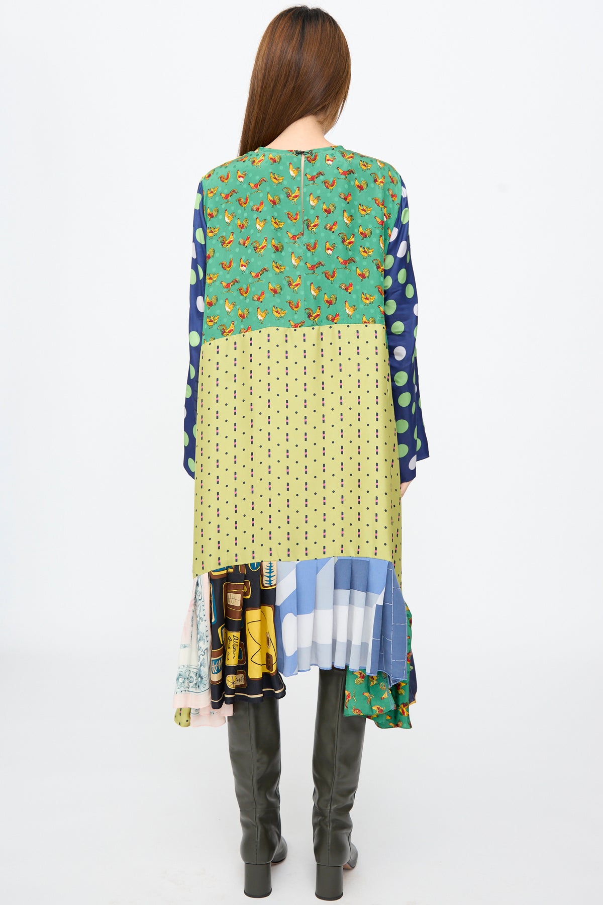 Back view of the Bettina Bakdal Hannah Dress in Friederike Dicker, a knee-length patchwork dress featuring green chicken-print, yellow polka-dot, and colorful patterns, worn with knee-high dark boots.