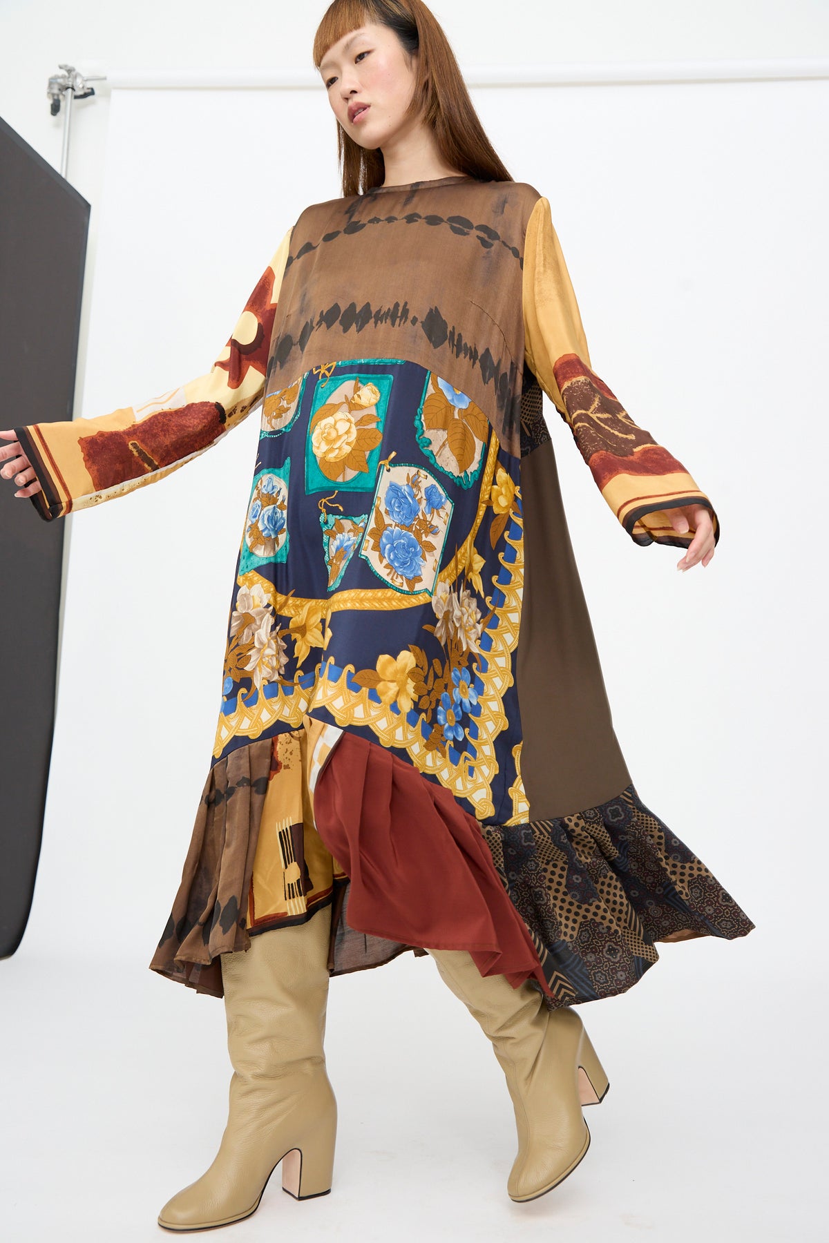 A person wearing the Bettina Bakdal Hannah Dress in Ise Gropius, a knee-length, long-sleeved up-cycled silk patchwork dress in brown, blue, and gold, paired with beige knee-high boots.