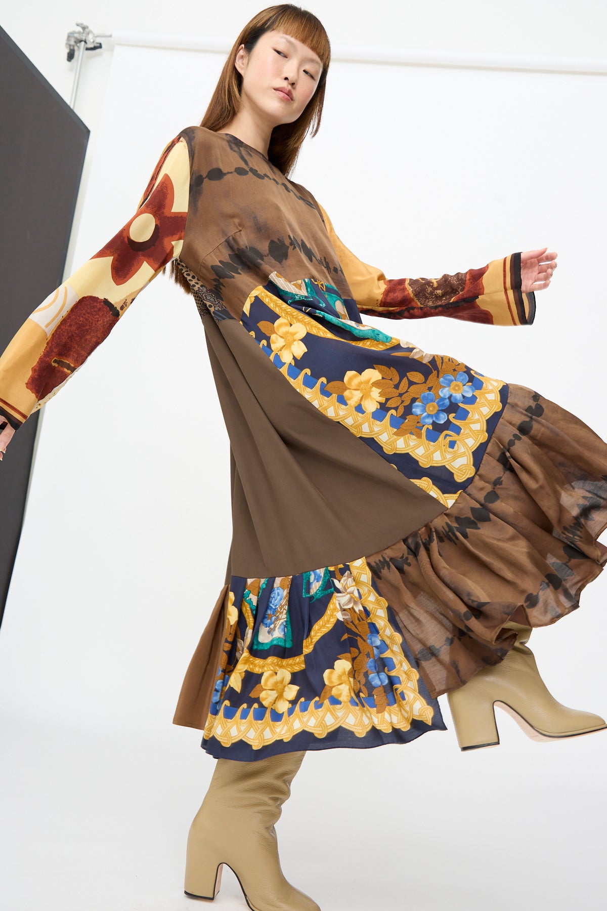 A model wears the Bettina Bakdal Hannah Dress in Ise Gropius, a long-sleeved, brown up-cycled silk dress featuring floral and geometric patchwork, a ruffled hem, and light-colored block-heeled boots.
