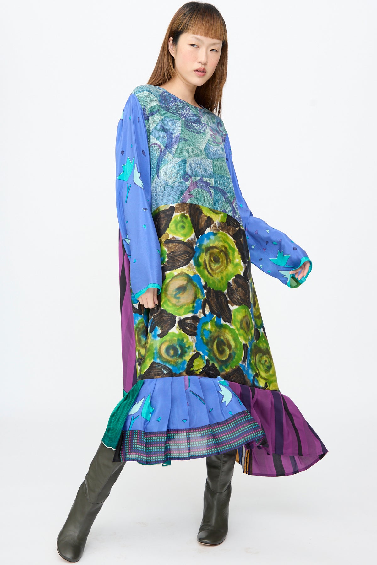 A person models the Bettina Bakdal Hannah Dress in Lucia Moholy, an up-cycled silk patchwork dress featuring a knee-length blend of blue, green, and purple floral patterns and dark knee-high boots.