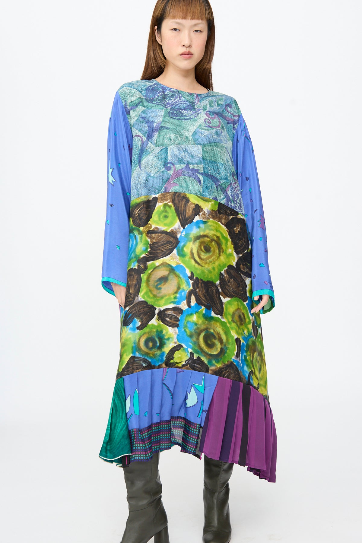 A person wears the Bettina Bakdal Hannah Dress in Lucia Moholy, a vintage silk patchwork dress with blue geometric patterns, large green floral motifs, and a pleated, multicolored hem.