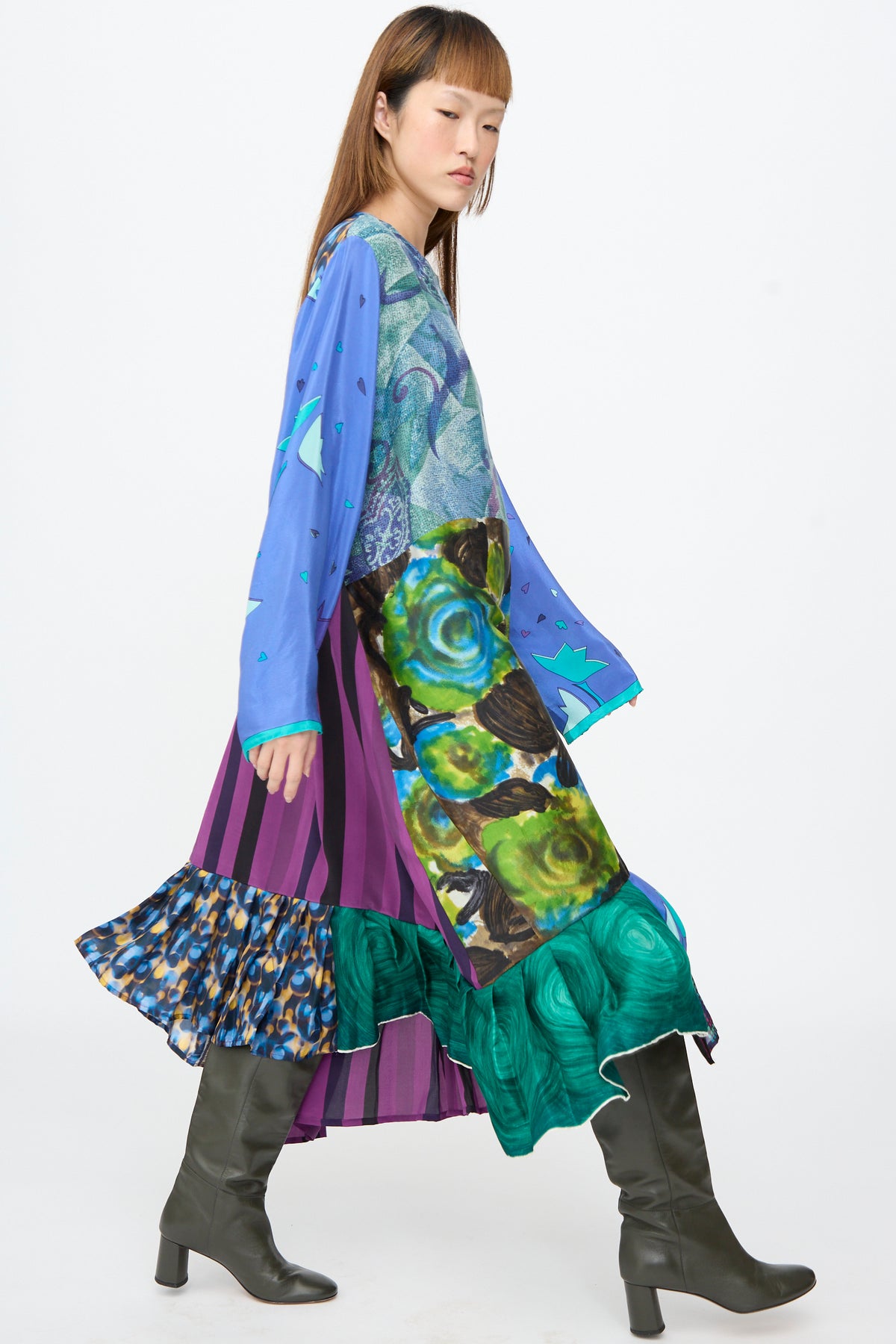 A person wears a blue, purple, and green vintage silk patchwork Hannah Dress in Lucia Moholy by Bettina Bakdal, paired with knee-length dark boots.