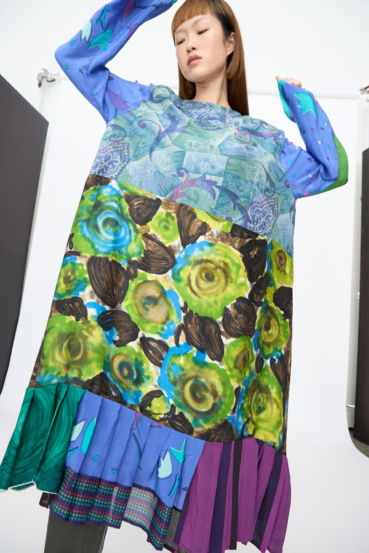 A person wearing the Bettina Bakdal Hannah Dress in Lucia Moholy, a knee-length, up-cycled silk patchwork dress featuring blue, green, and purple patterns with a pleated hem.