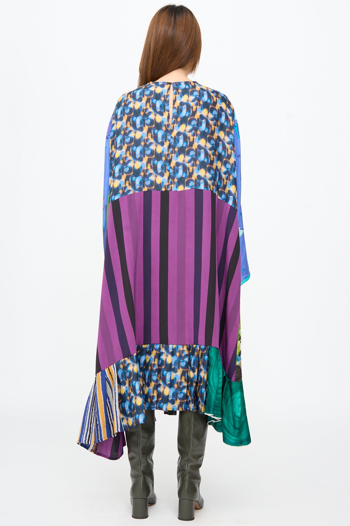 A woman seen from the back wearing a loose, knee-length Bettina Bakdal Hannah Dress in Lucia Moholy, featuring a patchwork of purple stripes, blue and yellow floral patterns, and green abstract shapes, paired with dark boots.