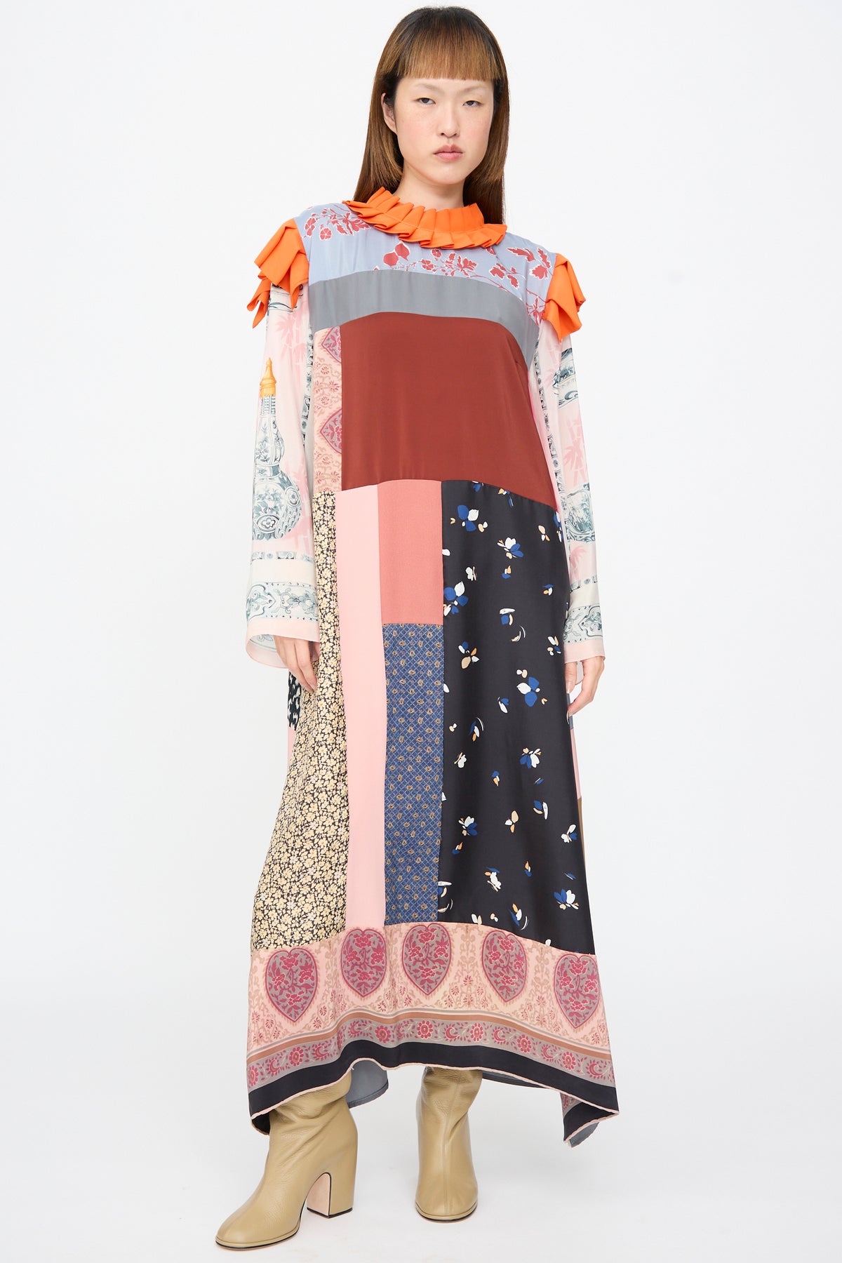 A person wears a Bettina Bakdal Ruffled Collar and Shoulder Dress in Grit Kallin, a long, up-cycled silk dress with a patchwork pattern, bright orange ruffled collar, and shoulder accents.