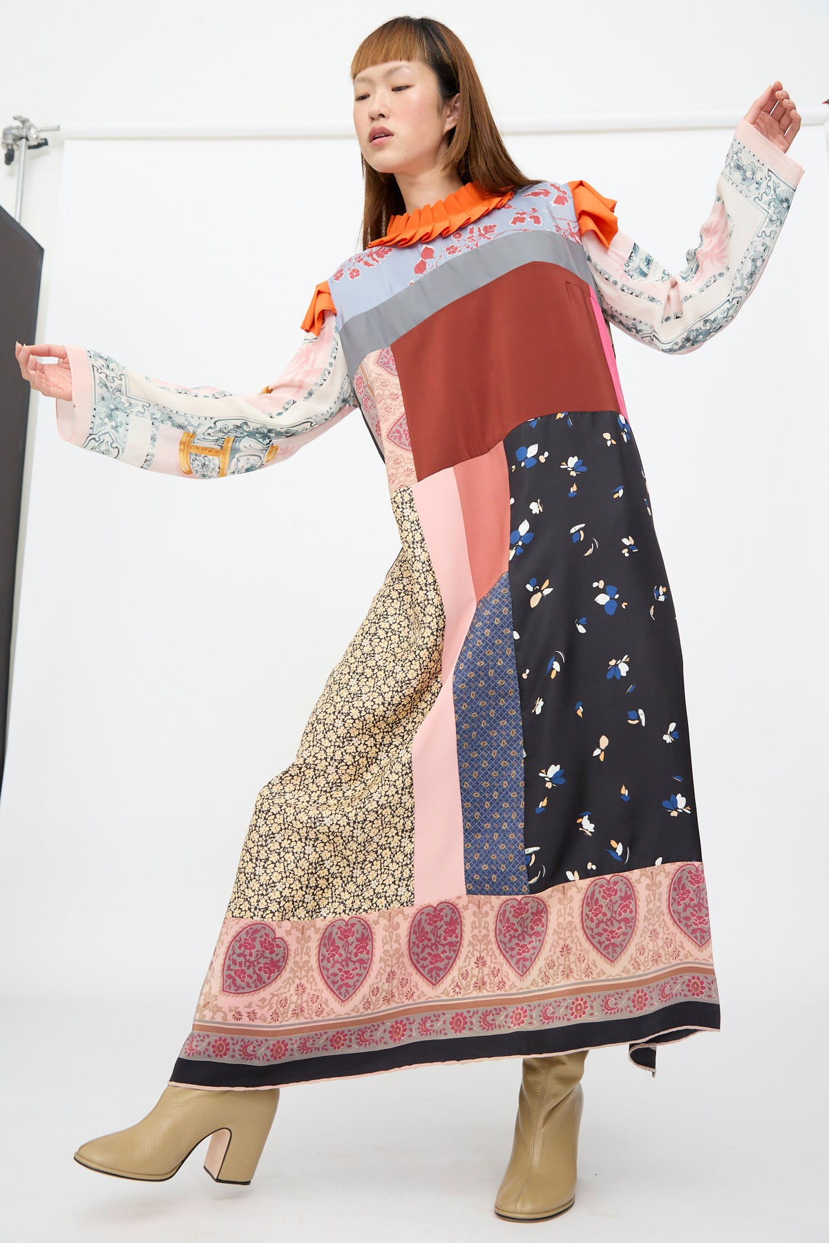 A person models the Bettina Bakdal Ruffled Collar and Shoulder Dress in Grit Kallin, a long-sleeved silk dress featuring an orange ruffled collar, floral and heart patterns, and beige block-heeled boots.