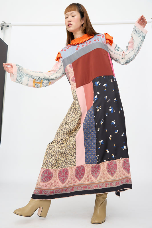A person models the Bettina Bakdal Ruffled Collar and Shoulder Dress in Grit Kallin, a long-sleeved silk dress featuring an orange ruffled collar, floral and heart patterns, and beige block-heeled boots.