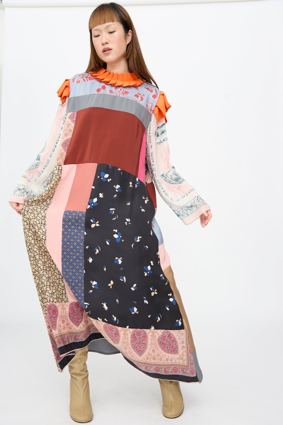 A model wears a long, loose-fit Bettina Bakdal Ruffled Collar and Shoulder Dress in Grit Kallin, featuring mixed patterned and solid-colored silk panels with a bright orange ruffled collar and accents.