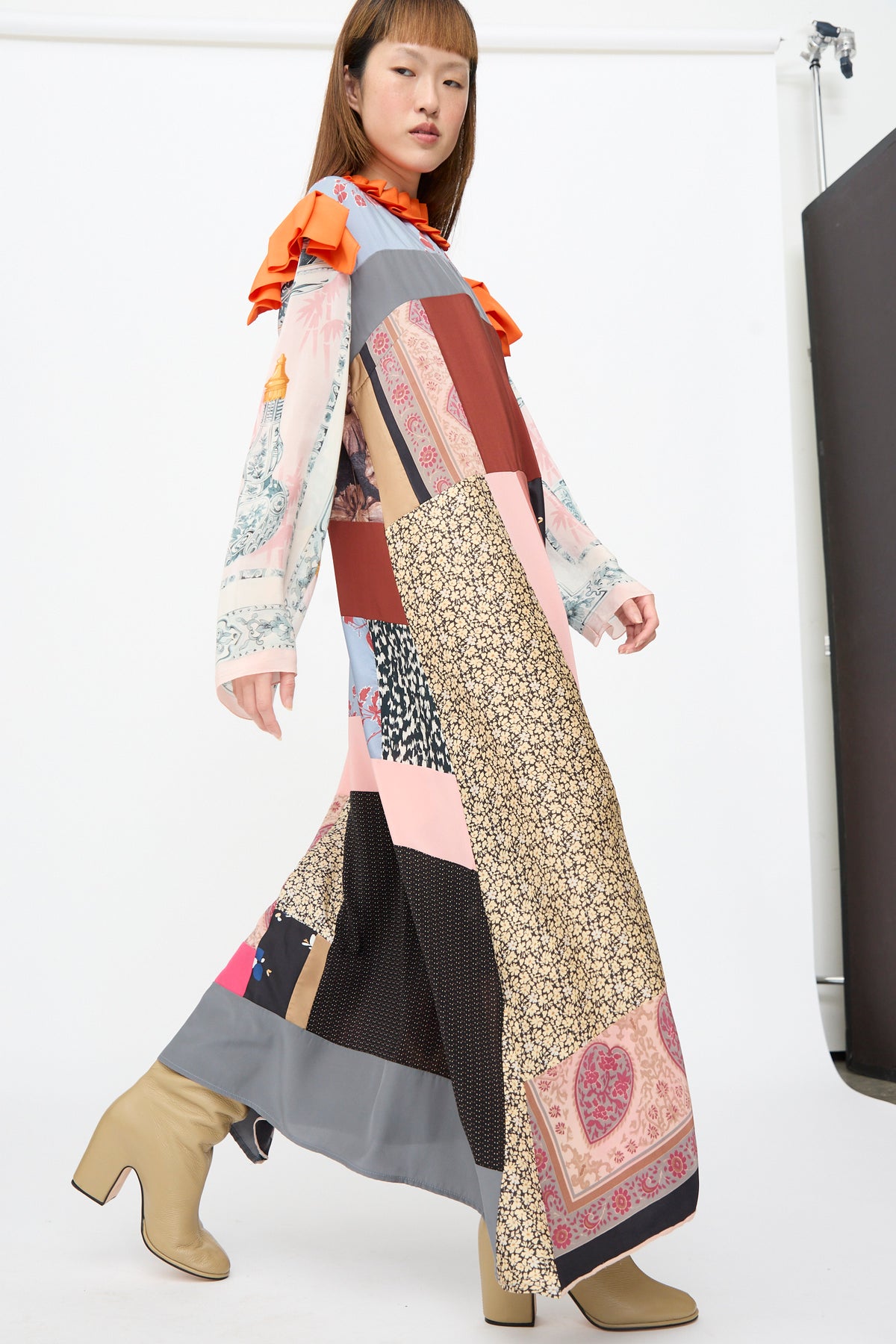 A person wearing a Bettina Bakdal Ruffled Collar and Shoulder Dress in Grit Kallin, paired with beige block-heeled boots.