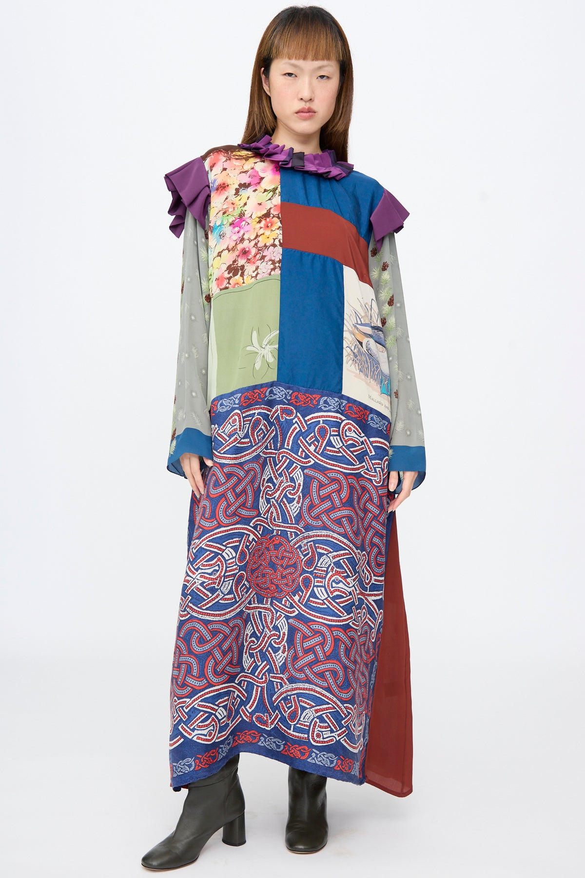 A person wears a Bettina Bakdal Ruffled Collar and Shoulder Dress in Ilse Fehli, a long-sleeve silk midi dress with a patchwork of floral and geometric patterns and a ruffled purple collar and shoulder.