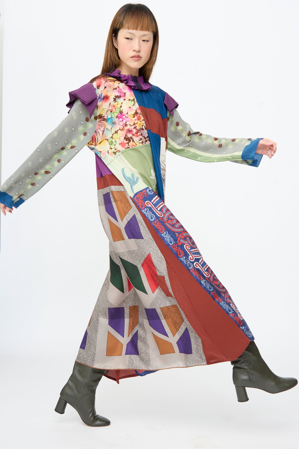 A person wearing a Bettina Bakdal Ruffled Collar and Shoulder Dress in Ilse Fehli, a long-sleeved, up-cycled silk midi dress with floral and geometric panels, styled with dark block-heeled boots.