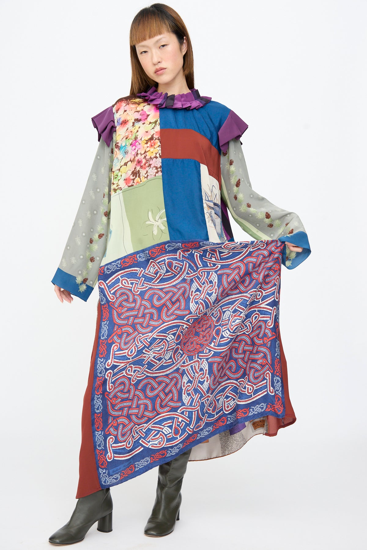 A person wearing a Bettina Bakdal Ruffled Collar and Shoulder Dress in Ilse Fehli, an up-cycled silk dress with floral patchwork, a ruffled collar, and a wide blue and red Celtic knot scarf panel.