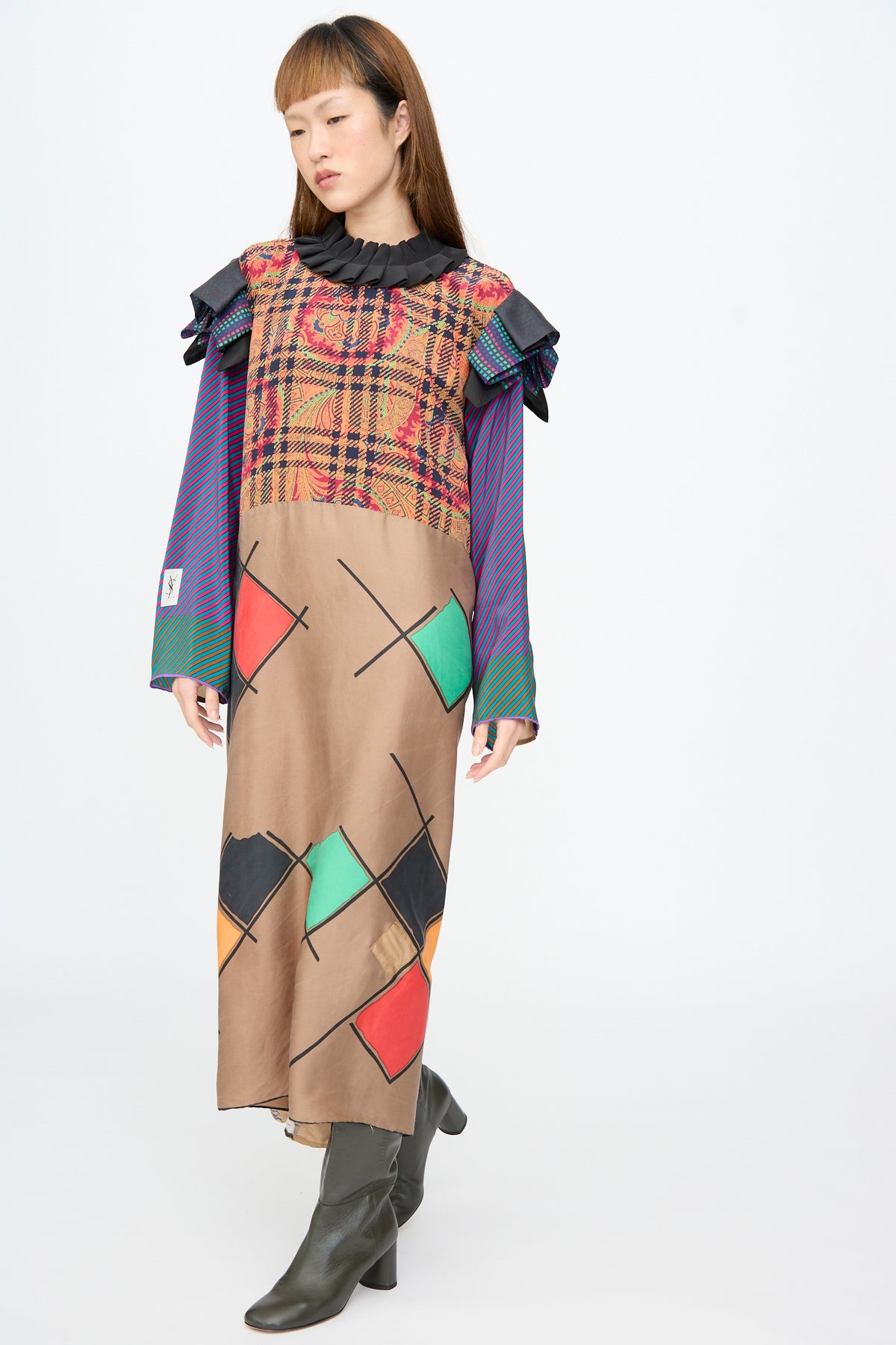 A model wears a Bettina Bakdal Ruffled Collar and Shoulder Dress in Marguerite Friedlaender, a midi-length patchwork dress with a ruffled black collar, multicolored plaid upper, and patterned tan lower.