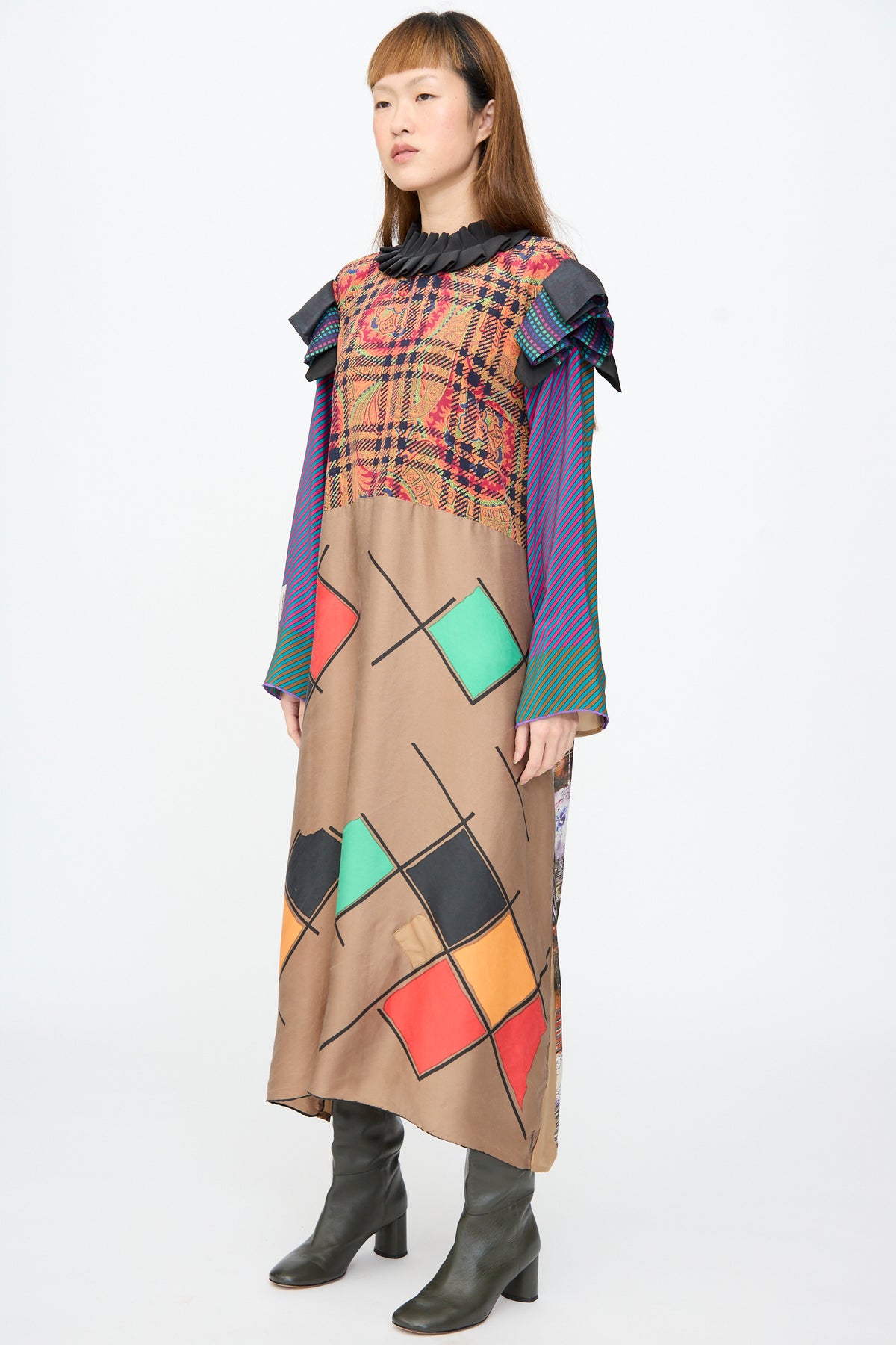 A person wears a Bettina Bakdal Ruffled Collar and Shoulder Dress in Marguerite Friedlaender, a long-sleeved up-cycled silk patchwork dress with a ruffled collar, plaid and striped panels, and colorful geometric squares, paired with dark heeled boots.