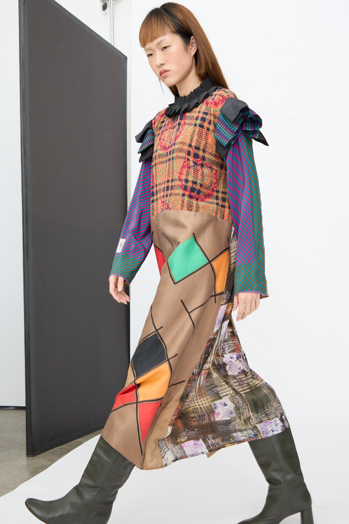 A model wears a Bettina Bakdal Ruffled Collar and Shoulder Dress in Marguerite Friedlaender, an up-cycled knee-length silk patchwork dress with mixed patterns, ruffles, and dark olive boots.