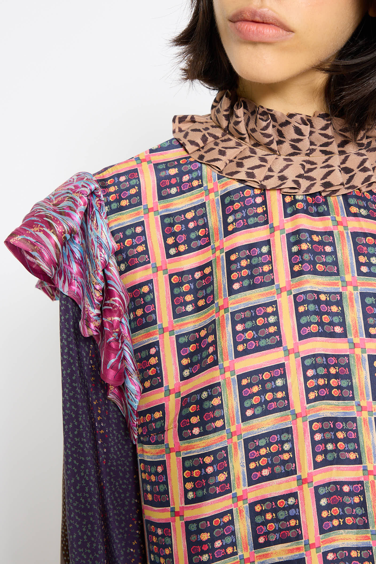 Close-up of a person wearing an up-cycled fashion piece—a colorful, patterned garment with geometric and floral designs. The outfit, originally a vintage silk dress by Bettina Bakdal called the Ruffled Collar and Shoulder Dress in Heather Blossom, features layered, textured fabric on one shoulder and a contrasting patterned collar.