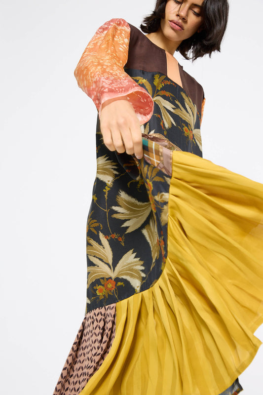 The person is wearing the Silk Hannah Dress in Forest Lunch by Bettina Bakdal, which features a relaxed fit and an intricate blend of floral and geometric designs crafted from up-cycled vintage silk, complete with a yellow pleated skirt section.