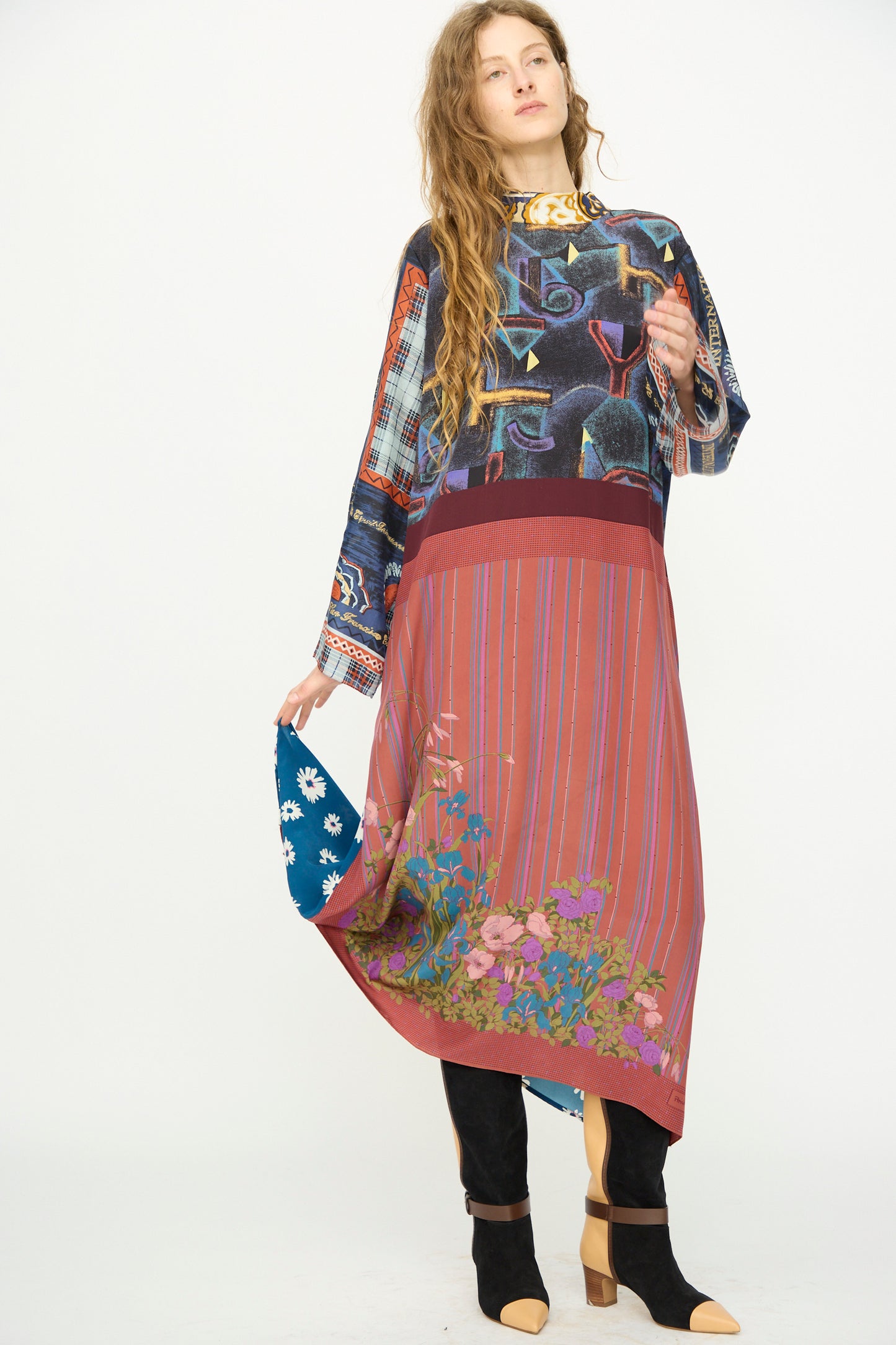 A woman stands against a white background wearing the Bettina Bakdal Classic Mandarin Collar Vintage Scarves Dress in Myriam Manuckiam, paired with black boots and holding a blue floral handkerchief.