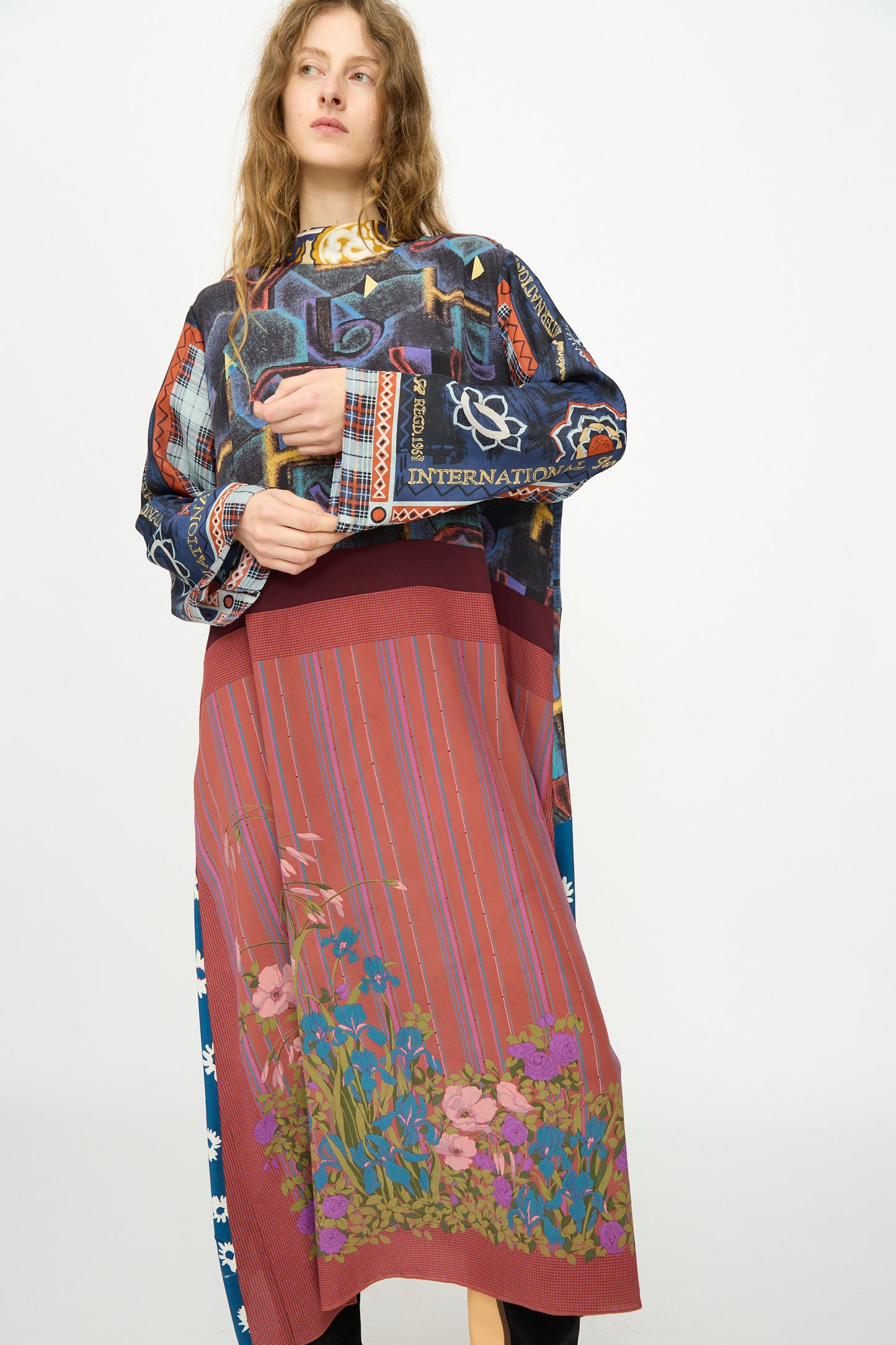 A woman with long wavy hair wears the Bettina Bakdal Classic Mandarin Collar Vintage Scarves Dress in Myriam Manuckiam, featuring a multicolored top, striped skirt, and floral hem, against a plain white background.