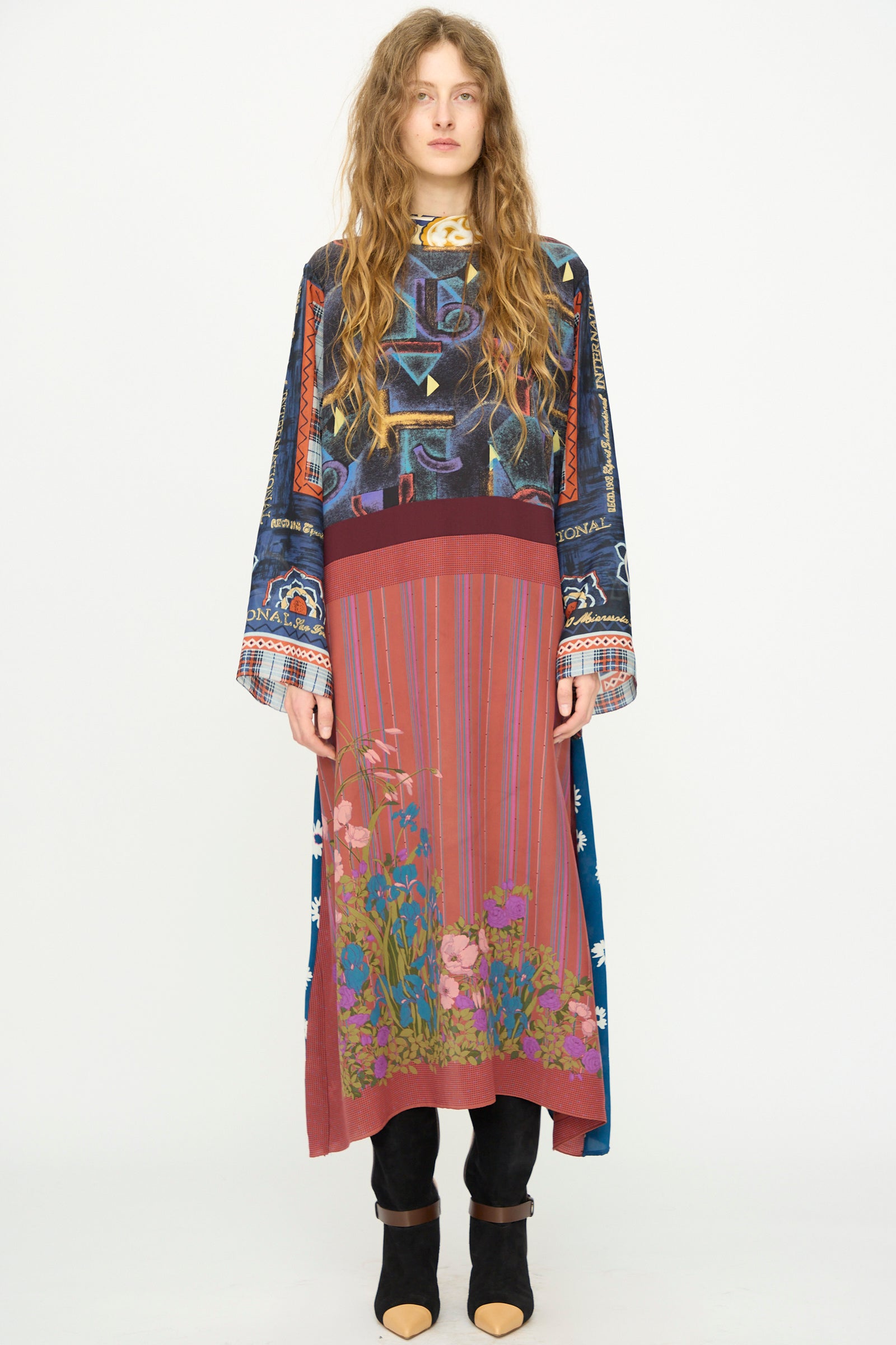 A woman with long wavy hair wears the Bettina Bakdal Classic Mandarin Collar Vintage Scarves Dress in Myriam Manuckiam, paired with black heeled shoes, standing against a plain white background.