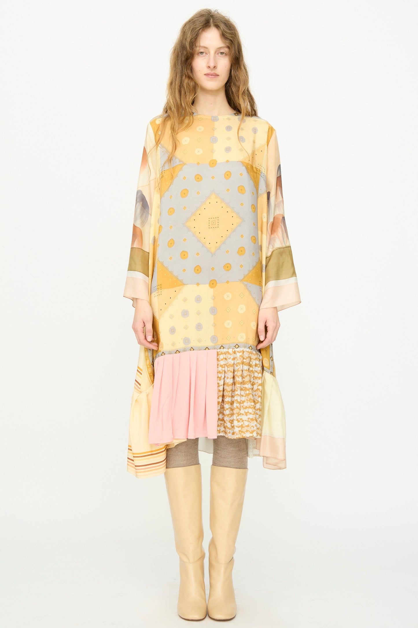 A person stands facing forward in the Bettina Bakdal Hannah Silk Dress in Pius Pahl, featuring yellow and gray vintage silk with ruffled hem, paired with beige knee-high boots against a plain white background.