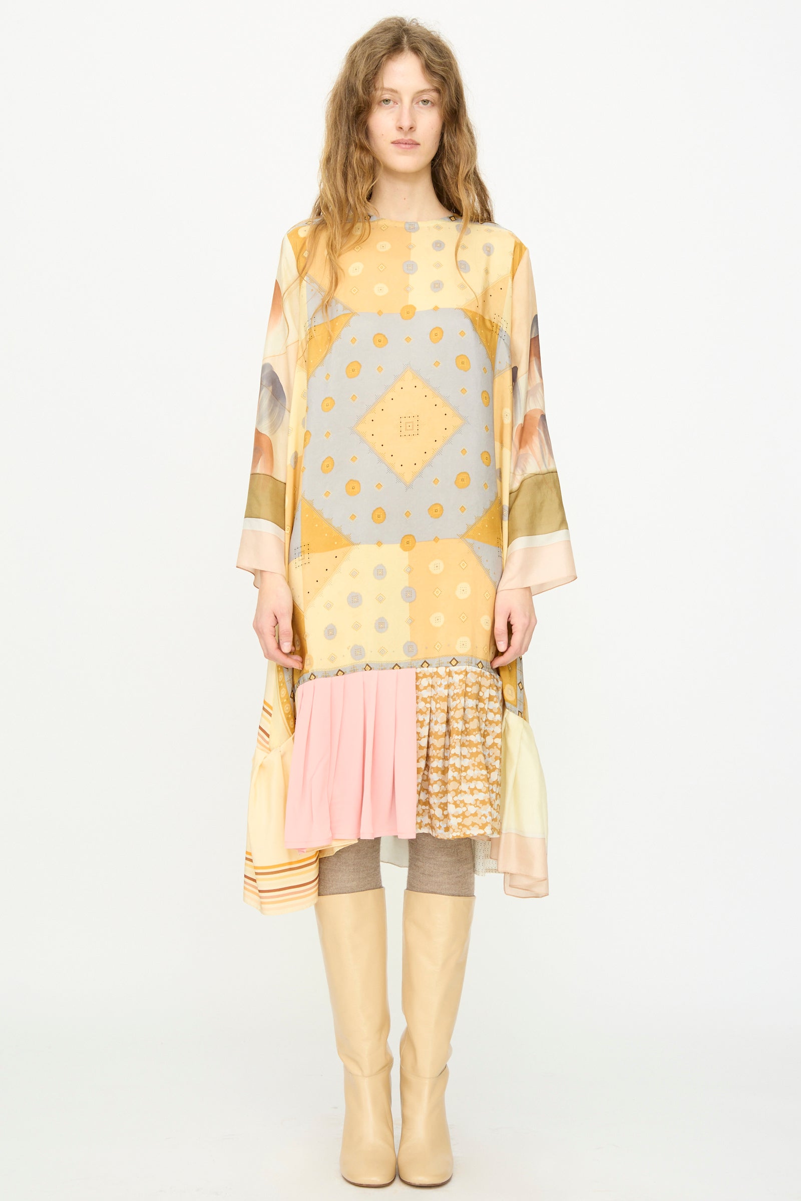A person stands facing forward in the Bettina Bakdal Hannah Silk Dress in Pius Pahl, featuring yellow and gray vintage silk with ruffled hem, paired with beige knee-high boots against a plain white background.