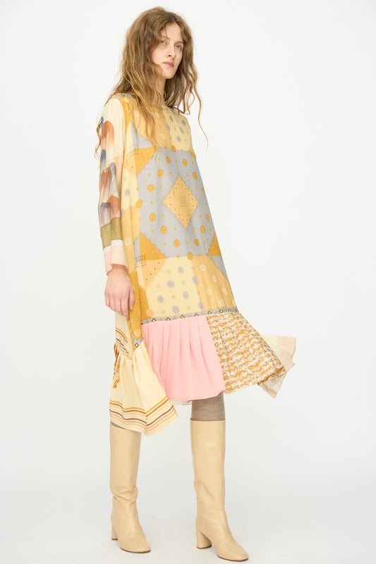 A woman wears the Hannah Silk Dress in Pius Pahl by Bettina Bakdal, featuring loose vintage silk with patchwork geometric and floral patterns, light pink and yellow pleats, paired with knee-high beige boots and a matching handbag.
