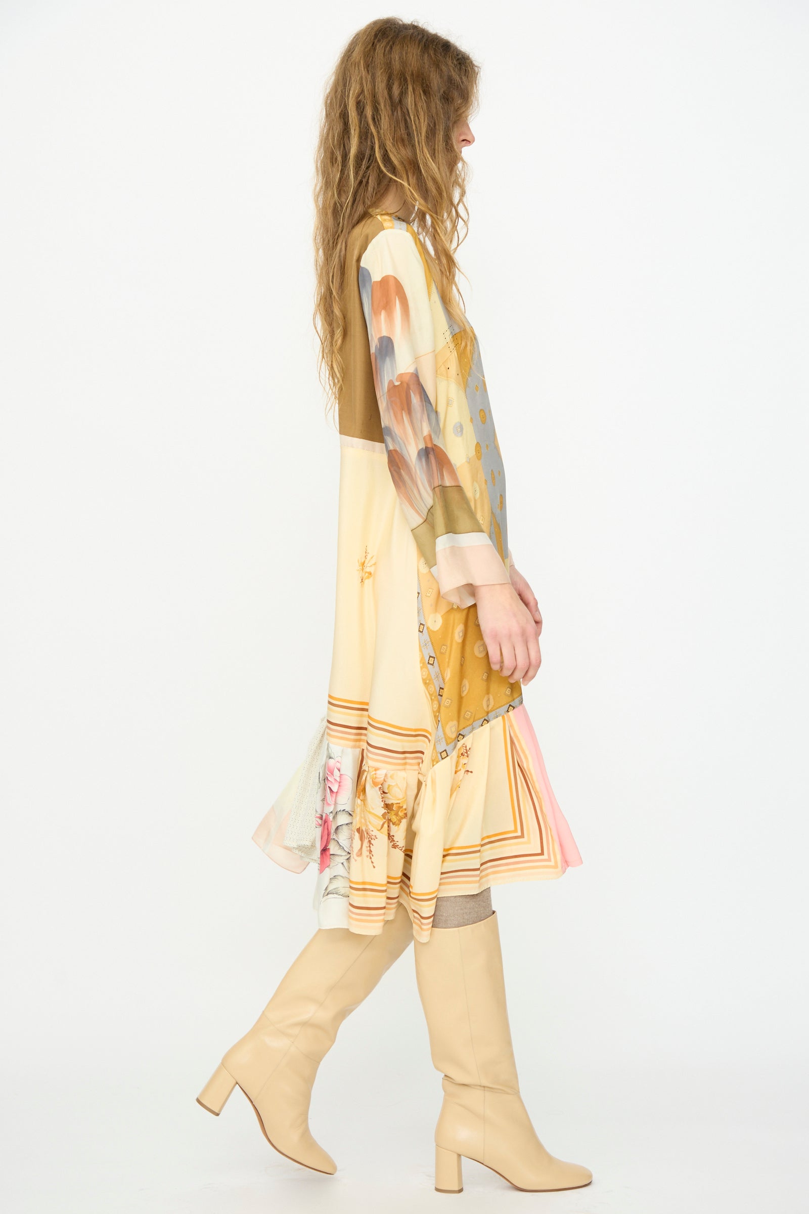 A woman with long wavy hair, wearing the Bettina Bakdal Hannah Silk Dress in Pius Pahl and light beige knee-high boots, stands in profile against a plain white background.