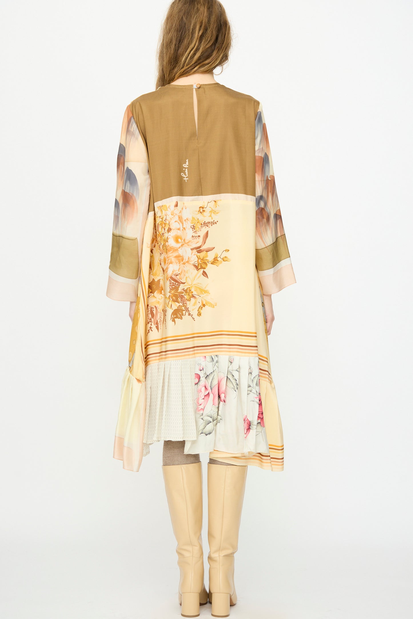 A person faces away, wearing Bettina Bakdal's Hannah Silk Dress in Pius Pahl—an up-cycled, knee-length dress with mixed floral and geometric patterns and long sleeves, styled with tall beige boots.