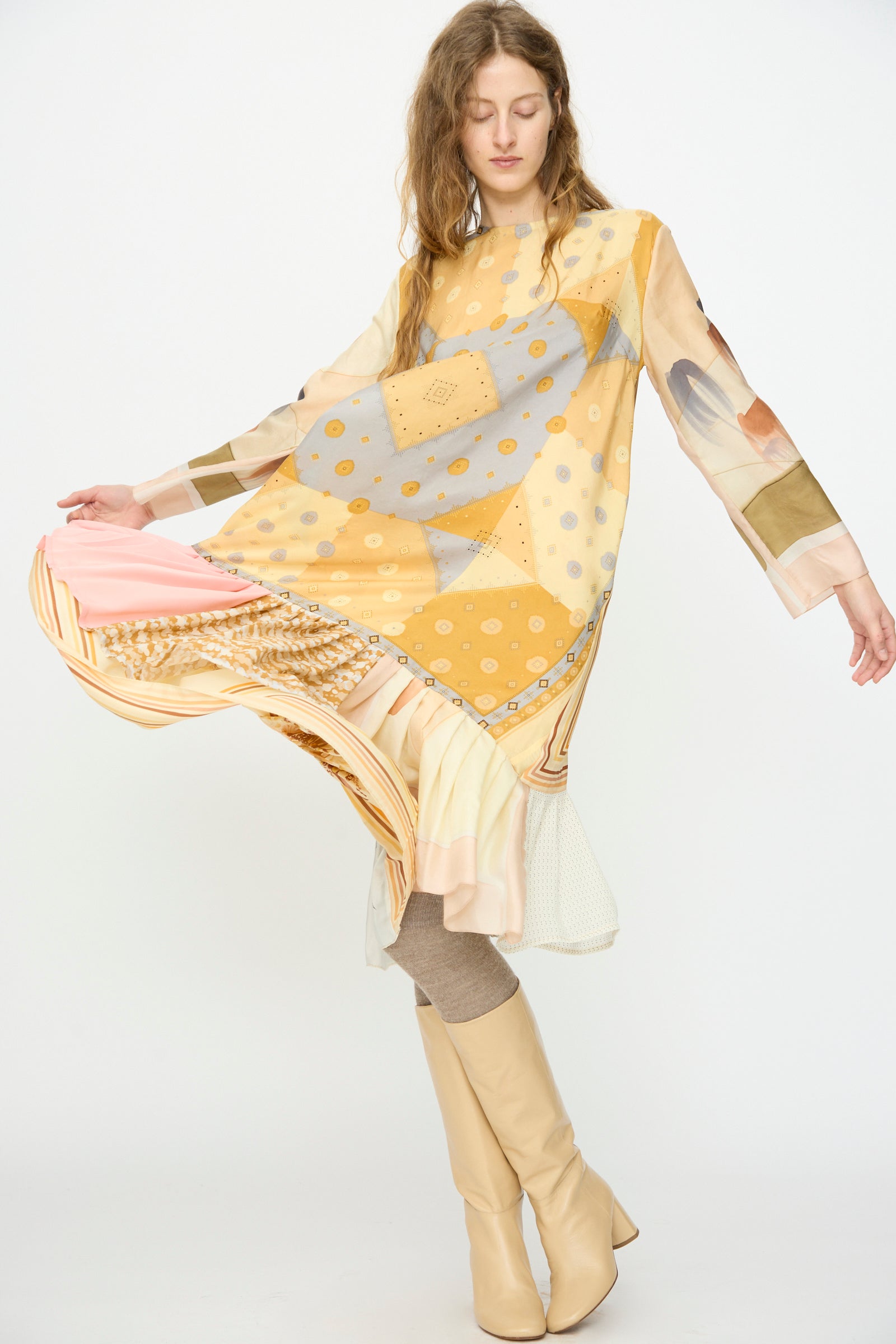 Someone wearing the Bettina Bakdal Hannah Silk Dress in Pius Pahl, featuring loose long sleeves and yellow patchwork, pairs it with beige boots and taupe tights while holding out part of the dress to the side.