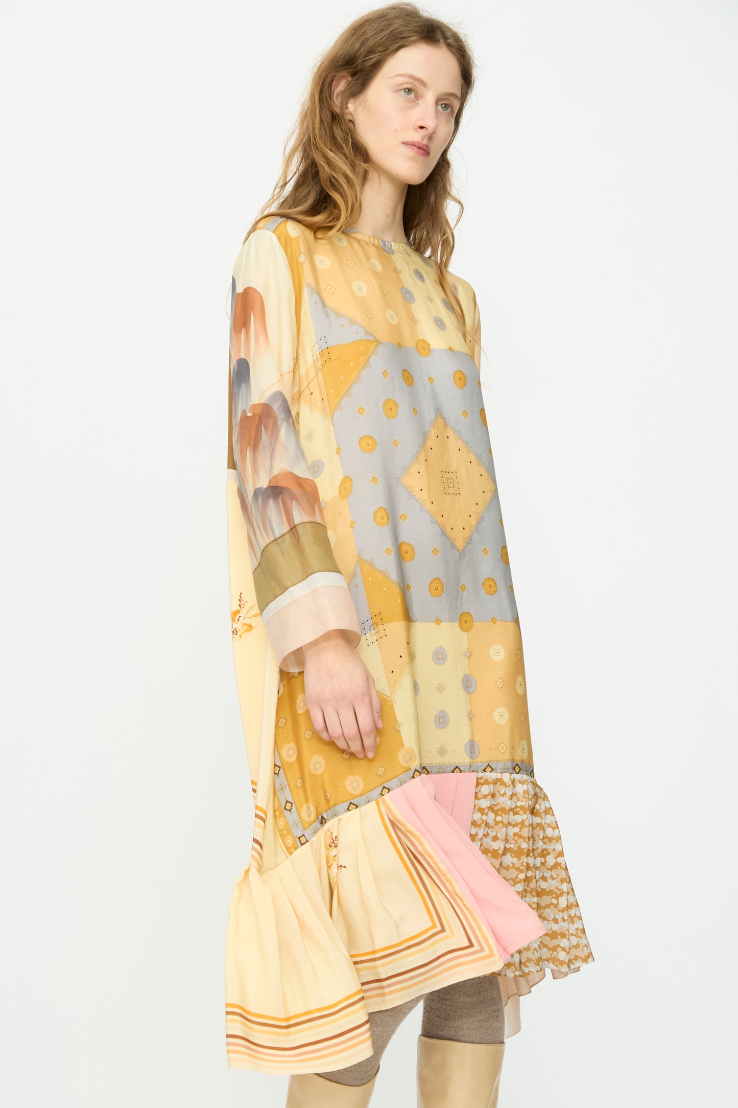 A woman wears the Bettina Bakdal Hannah Silk Dress in Pius Pahl, a loose, long-sleeved up-cycled silk dress with a patchwork pattern in gold, beige, and pastels and a ruffled hem inspired by vintage silk scarves.