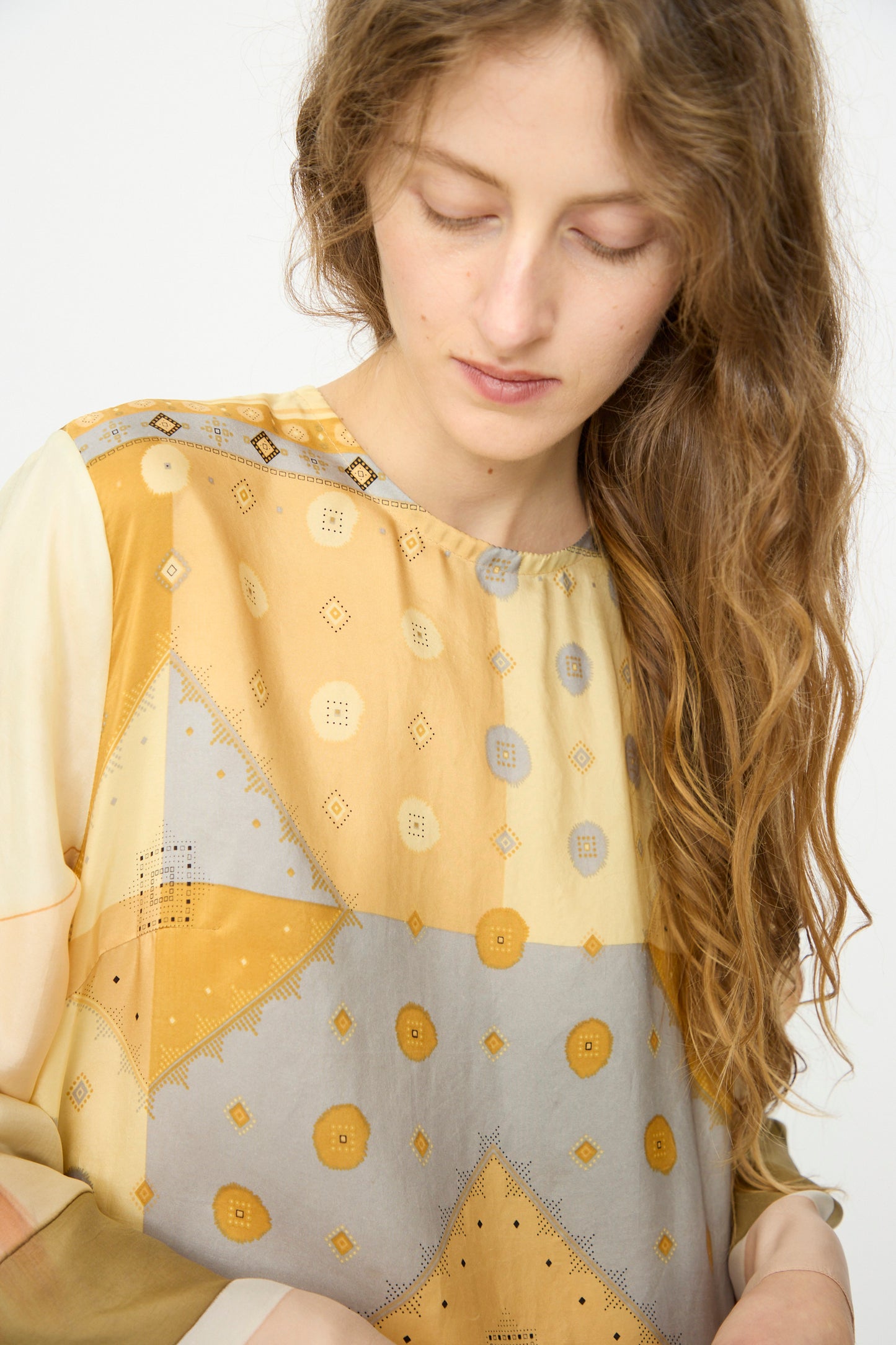 A woman with wavy brown hair looks down, wearing the Bettina Bakdal Hannah Silk Dress in Pius Pahl, a loose, patterned yellow and gray up-cycled silk dress, set against a plain light background.